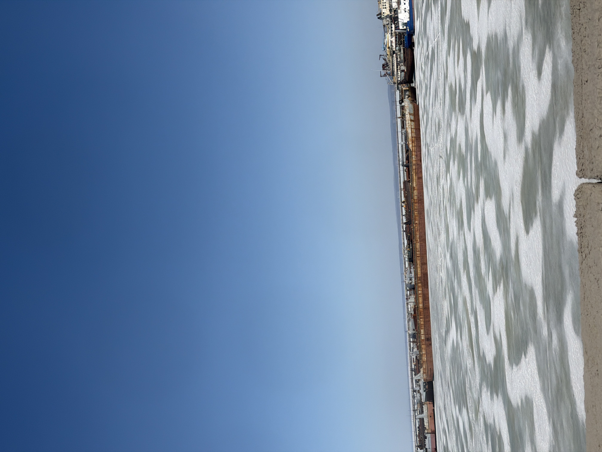 The image shows a view from what appears to be a high vantage point in the city of Chaikovsky, Russia. The right side of the image captures a section of the Votkinsk Reservoir. The water surface is covered in ice, displaying a pattern of light and shadows. Adjacent to the reservoir, the image reveals elements of a port or docking area with ships or industrial structures. The left side of the image is dominated by a clear, blue sky.