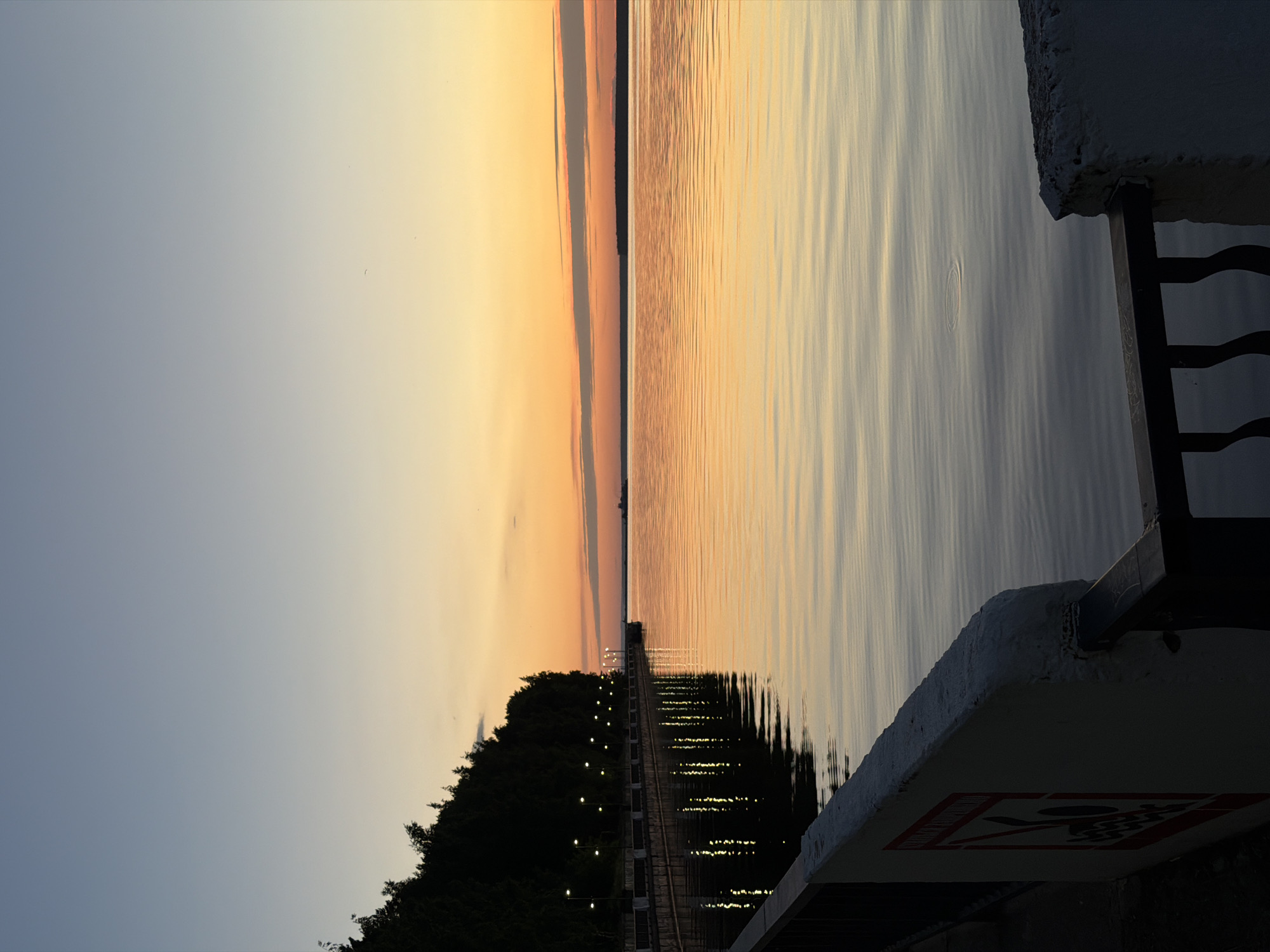 The image depicts a serene sunset over the Kama River in Chaikovsky, Russia. In the foreground, a concrete embankment with a black metal railing runs along the left side of the frame. A portion of a white wall with a warning sign featuring red and black graphics is visible on the far left. The embankment is lined with a row of illuminated lampposts, casting reflections on the calm water. To the left of the embankment, a dense line of dark trees forms a backdrop.
The middle ground is dominated by the Kama River, its surface reflecting the warm hues of the sunset. Gentle ripples are visible on the water. In the distance, the opposite bank of the river is marked by a dark silhouette of trees, partially obscured by the horizon.
The sky transitions from a clear, pale blue at the top of the frame to vibrant shades of orange and yellow near the horizon, indicating the setting sun. A few thin, elongated clouds are present in the orange band of the sky. The overall atmosphere is tranquil and peaceful. No people or animals are visible in the scene.