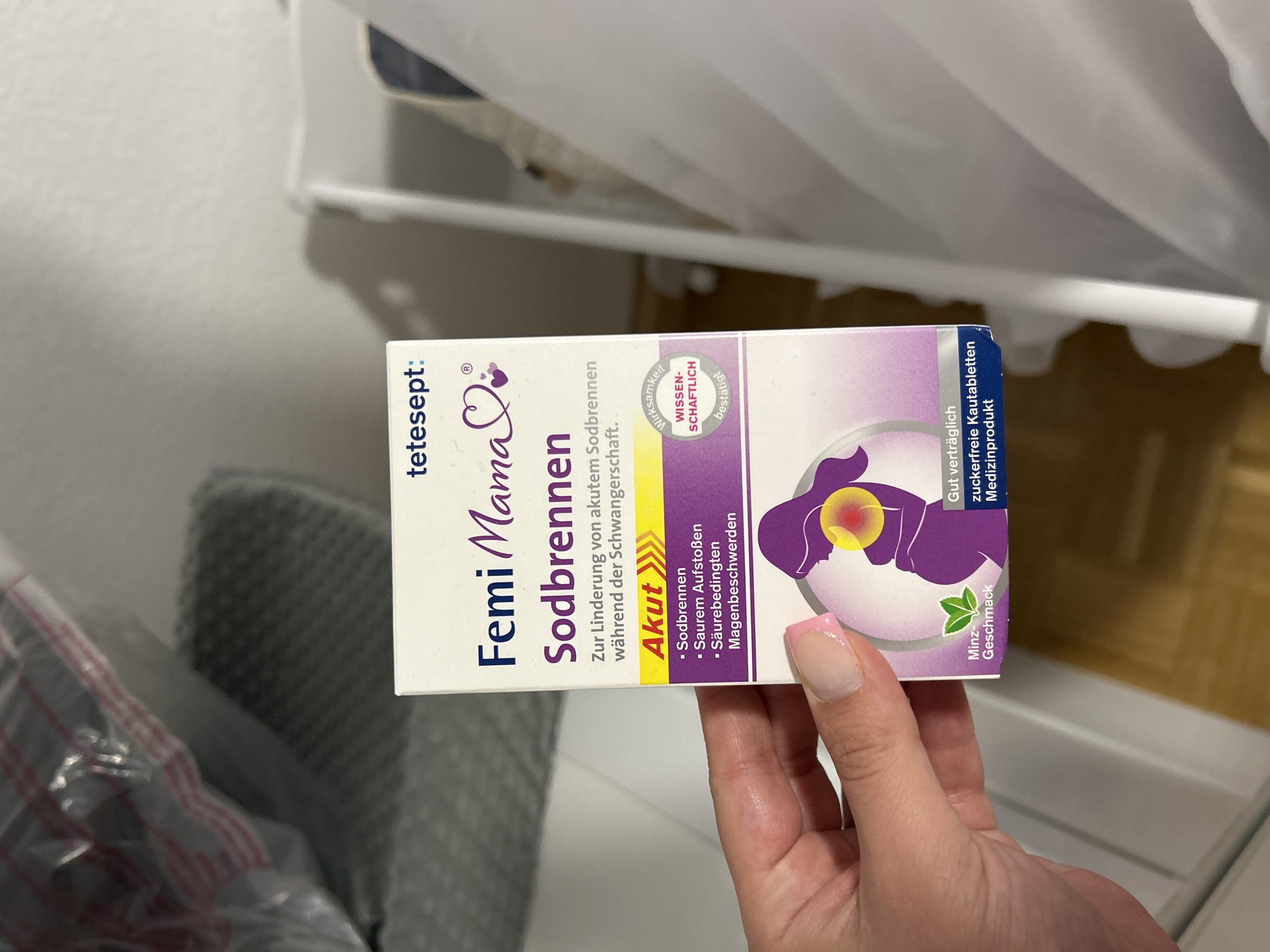 A close-up image depicts a fair-skinned hand holding a white and purple product box labeled "tetesept: Femi Mama Sodbrennen" in a residential indoor setting within Waldbröl, Germany. The box is oriented vertically, with its front face angled slightly towards the viewer, occupying the central and lower-right portion of the frame. The German text on the box identifies the product as "Sodbrennen" (heartburn) relief for "akutem Sodbrennen während der Schwangerschaft" (acute heartburn during pregnancy). Bullet points list targeted symptoms as "Sodbrennen," "Saurem Aufstoßen" (acid reflux), and "Säurebedingten Magenbeschwerden" (acid-related stomach complaints). A yellow strip with "Akut" (Acute) and arrows is visible. The box also states "Wirksamkeit WISSENSCHAFTLICH bestätigt" (Efficacy SCIENTIFICALLY confirmed), "Minz-Geschmack" (Mint-flavor), "Gut verträglich" (Well tolerated), and "Zuckerfreie Kautabletten Medizinprodukt" (Sugar-free chewable tablets Medical device). A purple silhouette of a pregnant woman with a stylized heartburn depiction (yellow and red glow) on her chest is prominent. The hand, exhibiting manicured fingernails with light pink tips, grasps the lower right side of the box, with the thumb visible on the front and fingers wrapping around the side.
The background is softly blurred. To the left, a clear plastic bag with some indistinct contents (possibly textiles) rests on a dark gray, textured surface, which might be a laundry basket or storage container. To the right, a white, slatted structure, consistent with the side of a baby crib or bassinet, is partially visible. The floor beneath is a light brown, possibly wood or laminate, and the walls are light-colored. The scene is illuminated by ambient indoor lighting.