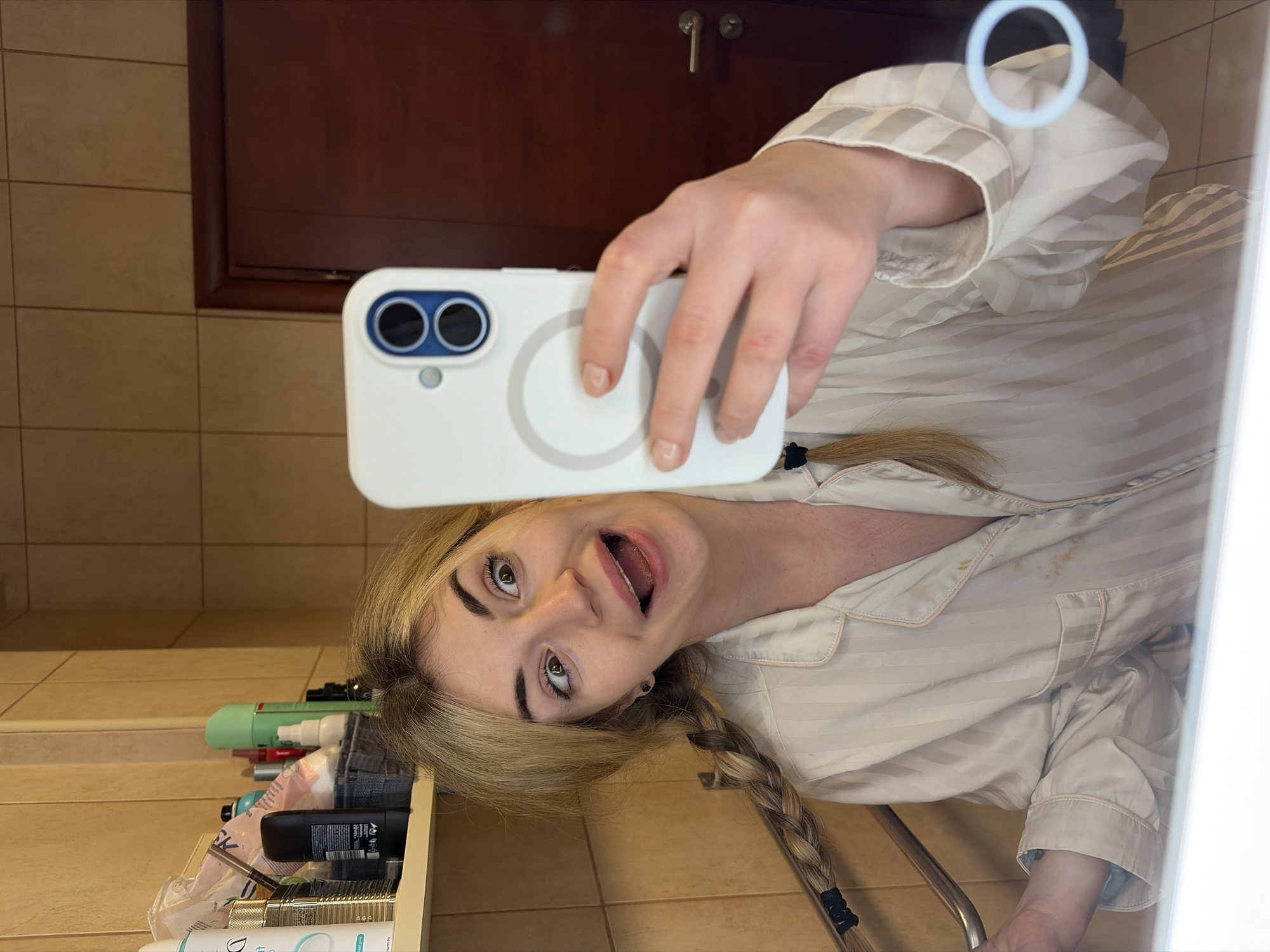 An adult female, reflected in a mirror, takes a selfie in a bathroom located in Budva, Montenegro. She has blonde hair styled in two braids and wears a light-toned, striped pajama top with pink piping, exhibiting minor stains on the chest area. Her expression is exaggerated, featuring wide, slightly crossed eyes and an open mouth. She holds a white smartphone with a visible MagSafe ring and dual blue-ringed camera lenses in her right hand. The bathroom interior includes light brown square wall tiles, a dark brown wooden door with a silver handle, a metal bar, and a shelf displaying various personal care products.