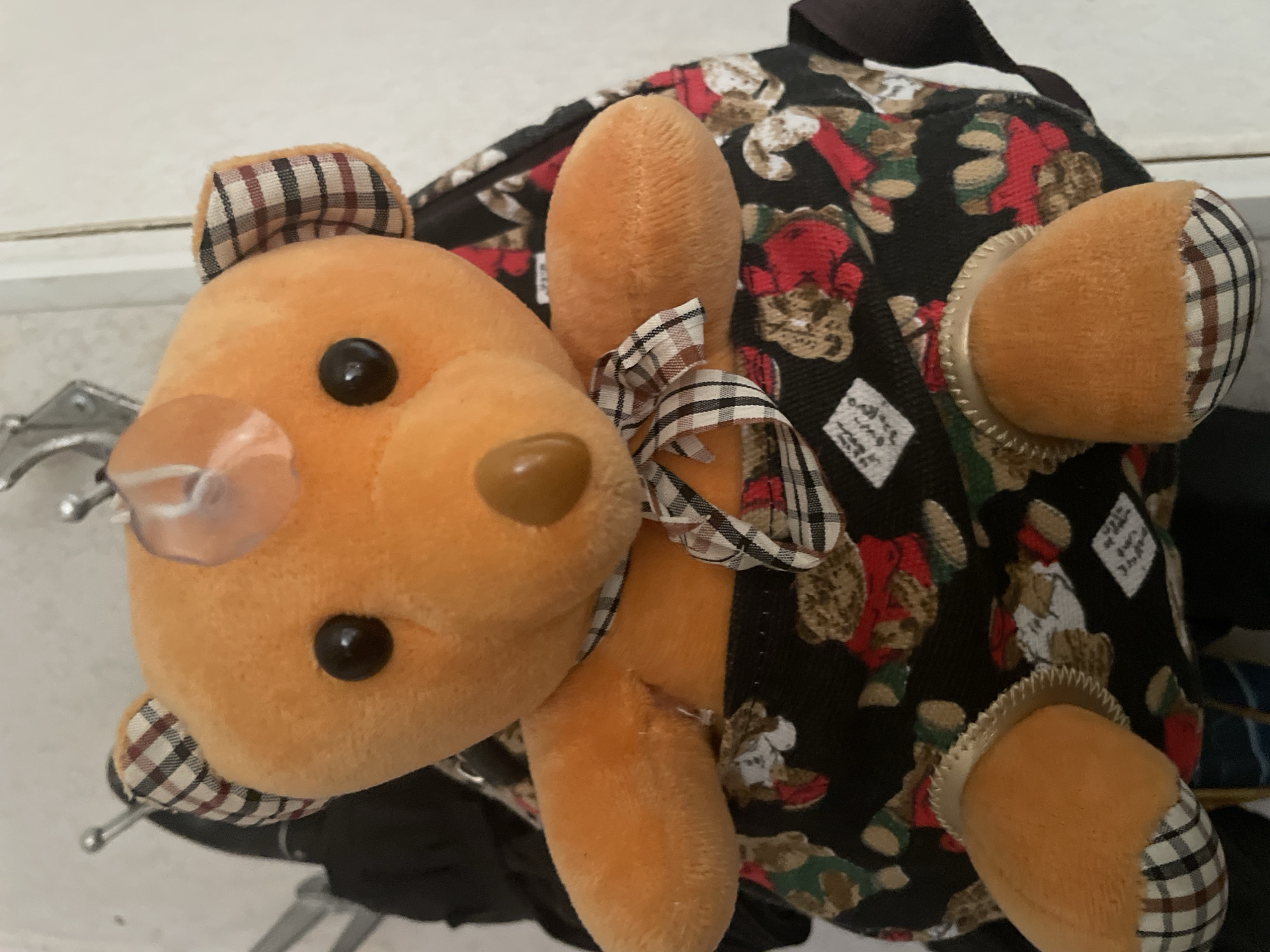 An orange plush teddy bear is displayed inside a black backpack with a repeating pattern of teddy bear graphics. The teddy bear has a plaid interior in its ears and a plaid ribbon tied around its neck. A clear suction cup is attached to the top of the teddy bear's head. The backpack is hanging on a hook, with a portion of another black object visible behind it. The background is a plain white wall.