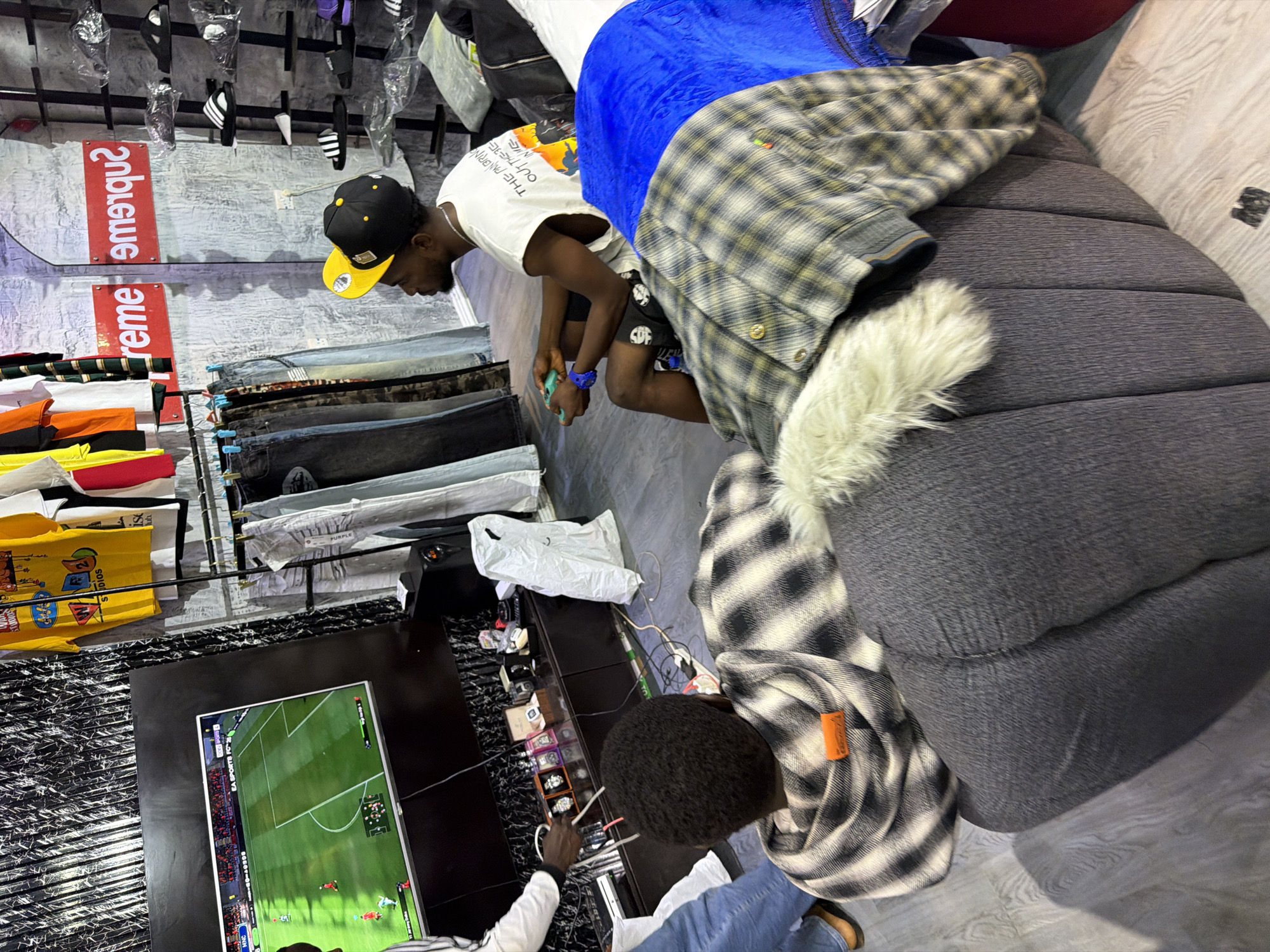 In a room that appears to be a store, two young Black men are present. One man, wearing a black and yellow baseball cap and a white t-shirt with text, is looking down and interacting with something out of frame. He is wearing dark shorts with a pattern. The second man, with dark, curly hair, is wearing a black and white plaid shirt and blue jeans. He is looking at a television screen that displays a soccer game.
The room is filled with various items, including clothing racks with t-shirts and jeans, and what appear to be shoes displayed on the wall. There are also some plush seating areas, one of which is a large, dark grey cushioned sofa. Several white plastic bags and other miscellaneous objects are scattered around. A television is mounted on a dark cabinet that displays an assortment of watches. Red and white "Supreme" logos are visible on the wall. The overall impression is of a retail environment, possibly a clothing store, with a relaxed atmosphere. The lighting suggests it could be daytime, and no specific weather conditions are evident. The presence of the soccer game on the television suggests leisure or entertainment might be a factor in the scene.
