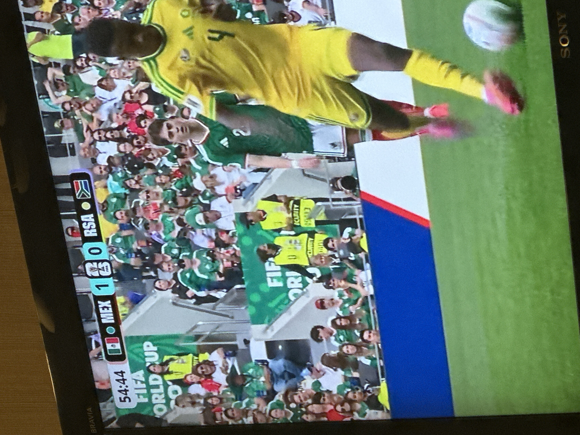 The image shows a televised soccer match between Mexico and South Africa. The score is displayed as 1-0 in favor of Mexico. The match is taking place in a stadium with a crowd of spectators visible in the background. The players are wearing their respective team uniforms. A player in a yellow jersey is kicking a soccer ball on the green field. The stadium appears to be modern, with seating areas and advertising boards. The overall lighting suggests it could be an indoor or nighttime event.