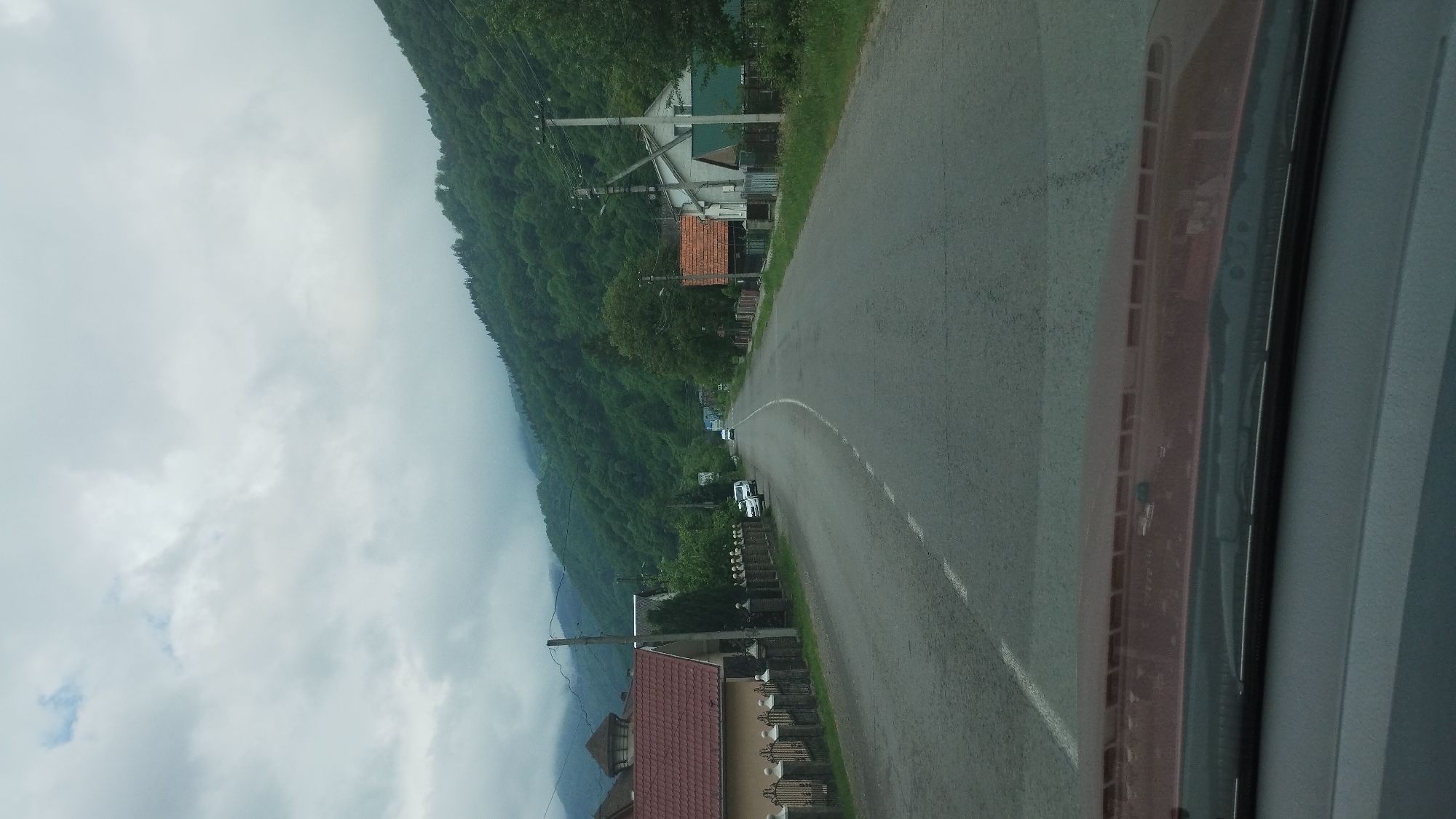 Captured from inside a vehicle, the image presents a road winding through the cityscape of Rakhiv, Ukraine. A mountain covered with lush, green forest dominates the left side of the frame, partially obscured by a cloudy sky. The road itself is gray asphalt with a white dividing line. Buildings with reddish-brown roofs are interspersed among the trees. The dashboard of the car is visible on the right side.