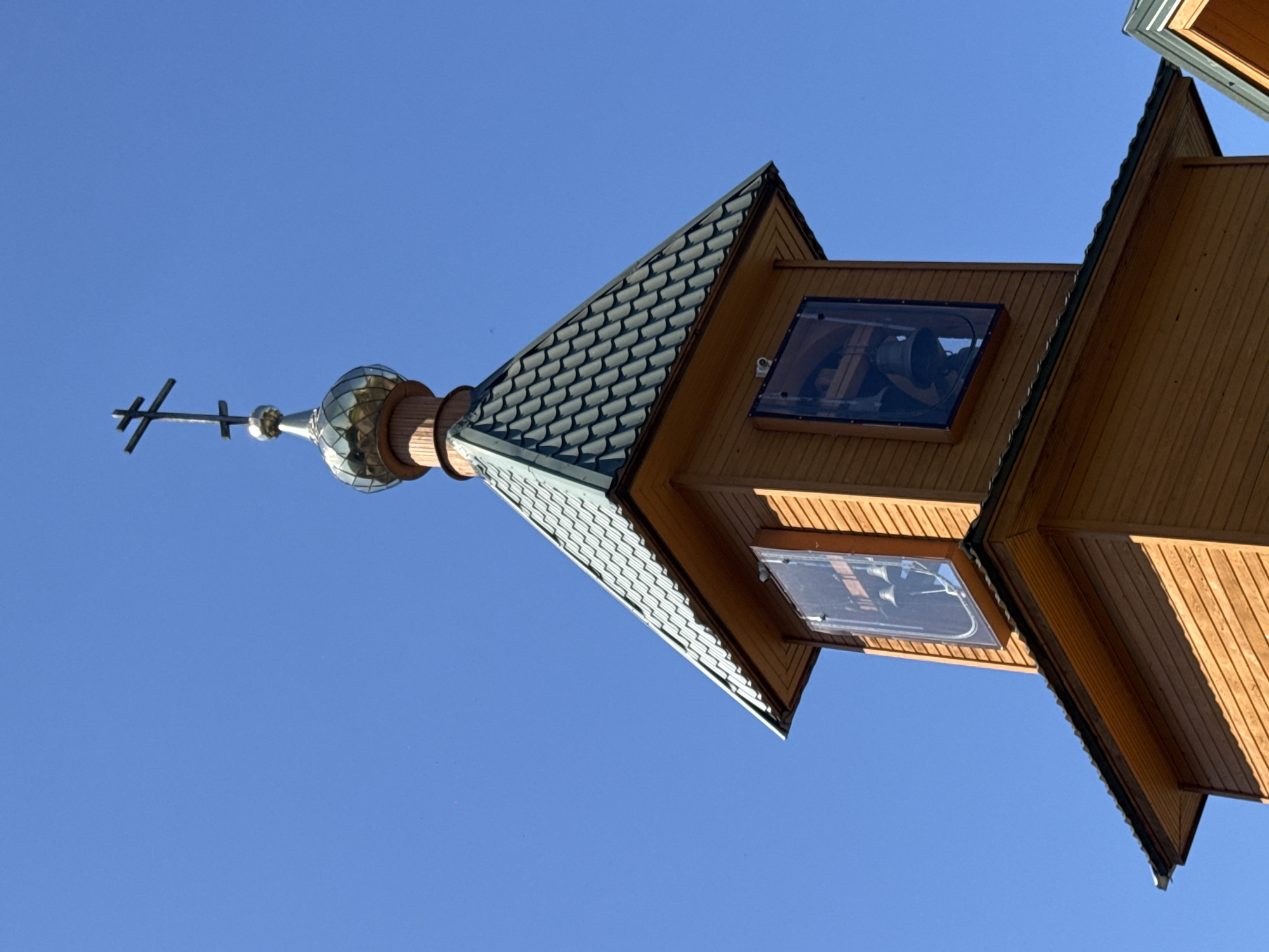 The image shows the top section of a wooden bell tower against a clear blue sky. The tower has a tiered, conical roof covered in dark shingles, topped with a golden dome and a three-barred cross. Two large, rectangular windows are visible on the tower's sides, each containing a bell. The wooden construction of the tower is evident with horizontal planks. The scene appears to be an exterior shot, likely of a church or monastery in Nizhny Novgorod, Russia, given the architectural style. There are no people or animals present in the image.