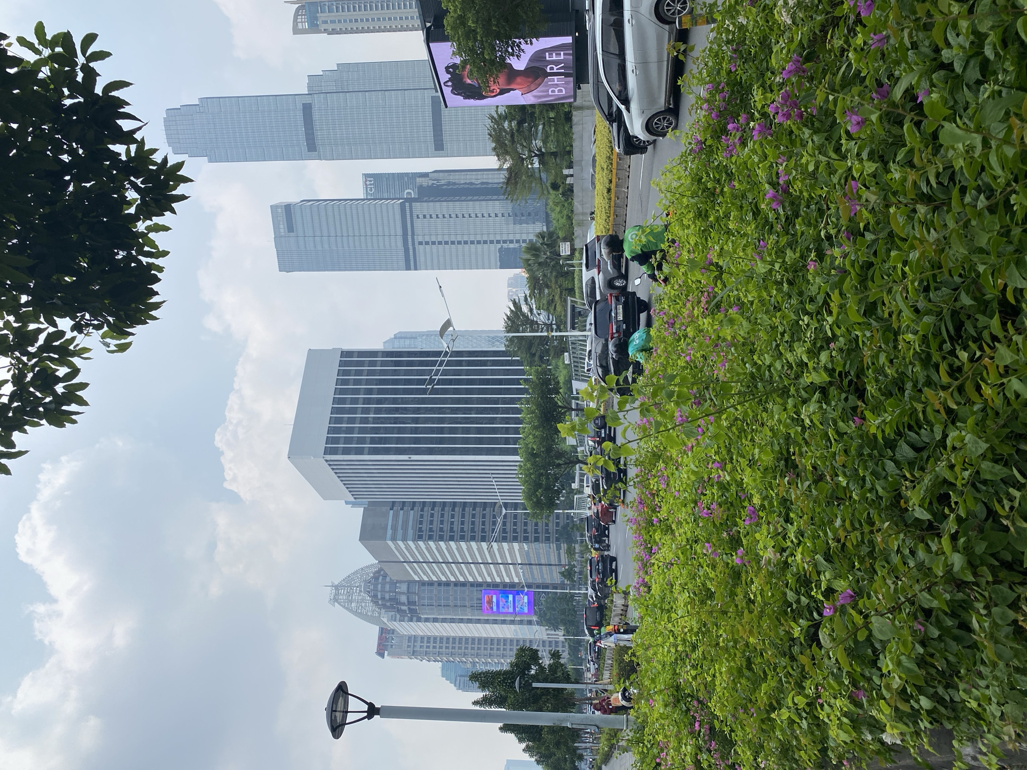 The image depicts a bustling street scene in South Jakarta, Indonesia, likely in the Kuningan or SCBD (Sudirman Central Business District) area, characterized by its modern skyscrapers and vibrant city life. In the foreground, a lush hedge of green foliage dotted with small purple flowers lines the street, creating a natural barrier. Beyond the greenery, a busy road is filled with a variety of vehicles, including cars, motorcycles, and a public transportation vehicle. Several people are visible, some walking along the sidewalk and others on motorcycles.
Dominating the background is a cluster of contemporary high-rise buildings, their glass facades reflecting the overcast sky. One prominent building displays a large digital billboard featuring a portrait of a person and the text "BHRE". A lamppost stands on the left side of the road, its design being a typical urban street fixture. The overall atmosphere suggests a dynamic urban environment with a mix of nature and architecture, typical of a major business and commercial district. The presence of what appear to be ride-sharing motorcycles, indicated by their bright green helmets and jackets, further points to a contemporary urban setting. The cloudy sky suggests a possibly humid day, common in tropical climates like Indonesia.