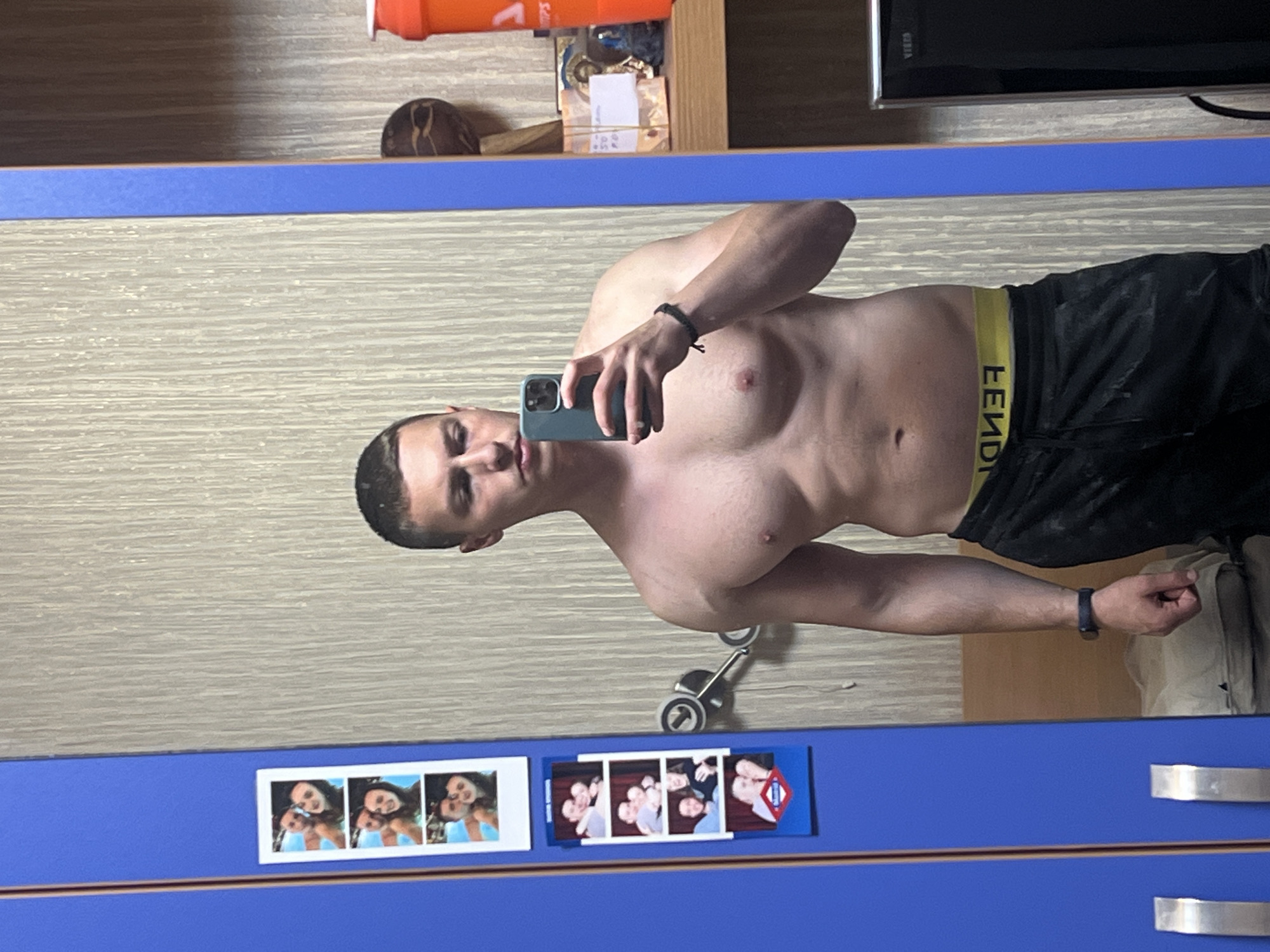 A shirtless man with short dark hair takes a selfie in a mirror. He wears dark athletic shorts with a yellow Fendi logo waistband. He has a dark bracelet on his left wrist. The mirror is mounted on a blue cabinet or dresser. Three small photo strips are adhered to the cabinet: one of three women, one of a group of people, and one that includes a symbol similar to the London Underground logo. A grey toy scooter is visible in the reflection. The background behind the man includes wooden shelves with various items, including a can of energy drink and a television. There is no information available to determine that the location is the city of Burgas, Bulgaria.