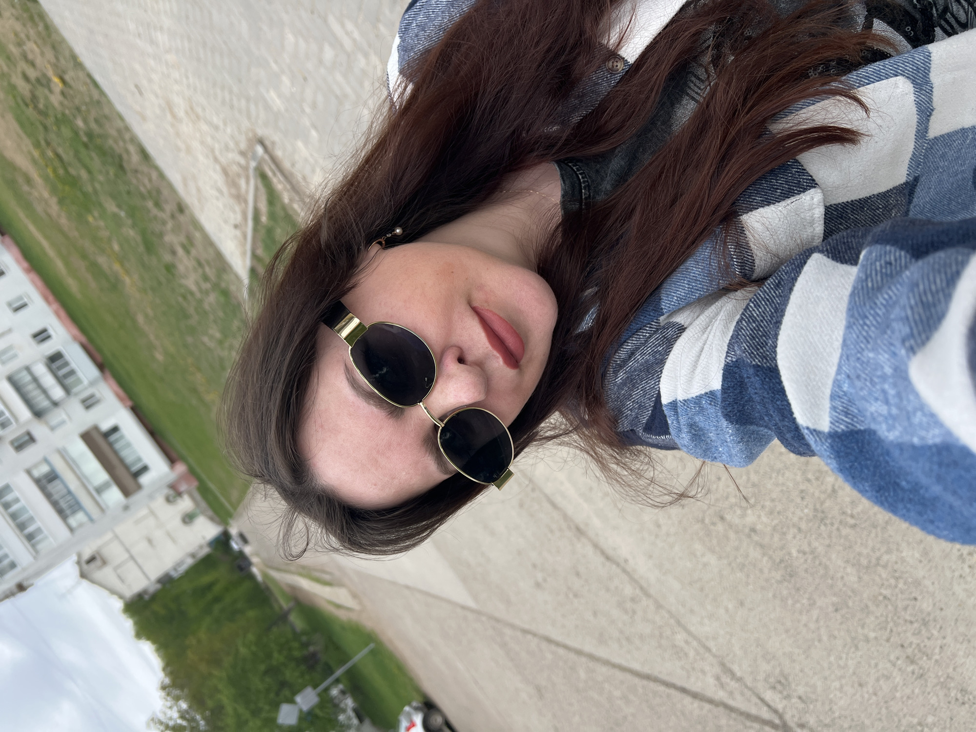 A selfie shot of a woman in Noviy, Russia. She is wearing sunglasses and a blue and white plaid shirt. Behind her is a sidewalk, grass, trees, and a building.