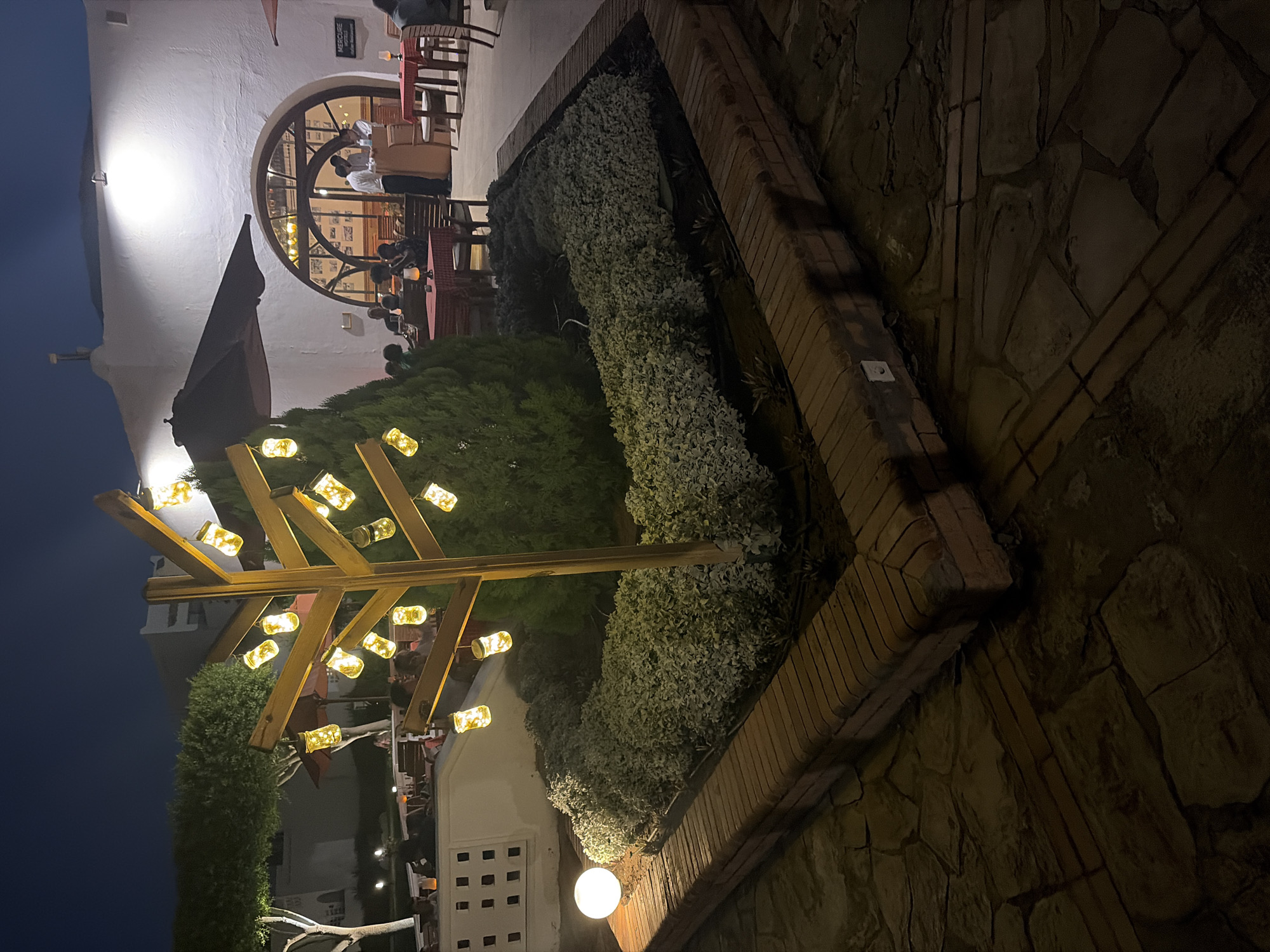 The image captures an outdoor seating area at night, likely a restaurant or cafe, in Hurghada, Egypt. In the foreground, a decorative wooden tree-like structure is illuminated by numerous small, glowing lanterns. Adjacent to it is a circular white lamp and a planter filled with lush green foliage, enclosed by a brick border. The background reveals the exterior of a white building with an arched entryway. Several tables and chairs are set up, with some patrons seated and staff attending to them. The overall ambiance is warm and inviting, with the lights creating a festive atmosphere.