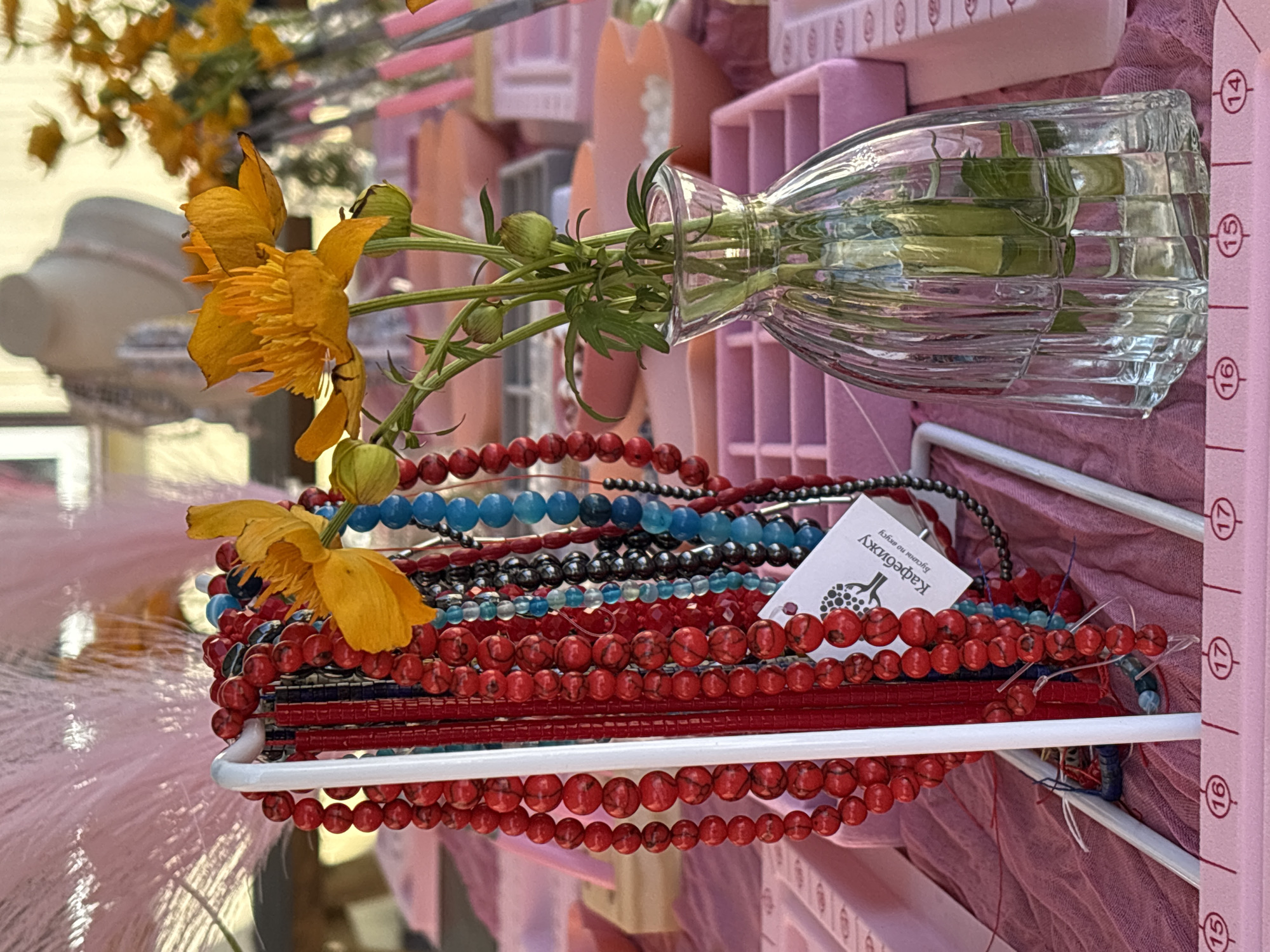 The image depicts a close-up of a stall at a market, possibly a craft fair. In the foreground, a display rack holds numerous strands of colorful beaded necklaces, predominantly in shades of red, blue, and black. A small white tag with Cyrillic text, "Кафебижу. Бусины по вкусу" (Kafebizhu. Beads to taste), is attached to one of the red necklaces. To the right, a clear glass vase contains a small bouquet of bright yellow flowers and green stems. The background reveals more market stalls, with displays of jewelry and various pink trays and boxes, suggesting an organized retail setup. A blurred image of a mannequin wearing a pearl necklace is visible in the far background. The overall setting suggests an outdoor or indoor market environment in Birobidzhan, Russia, focused on handmade crafts and accessories.
