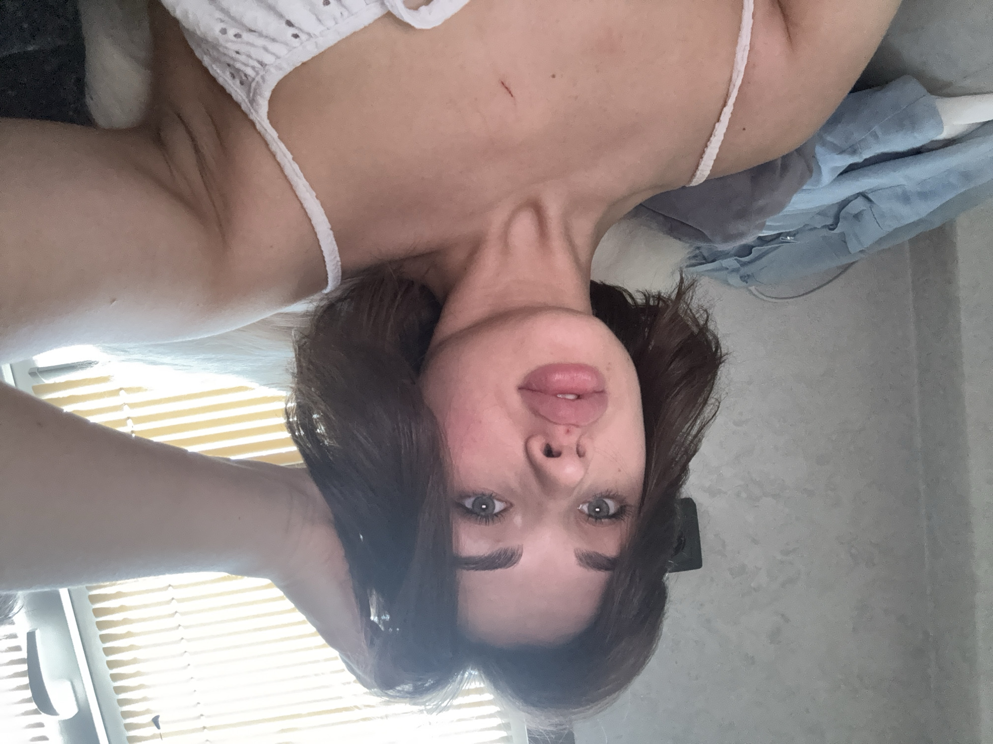 A young woman with brown hair and fair skin, lying down, wearing a white top, looking upwards with a slight frown. The background includes a window with blinds and a gray wall. The location is possibly in Лобаново, Russia.