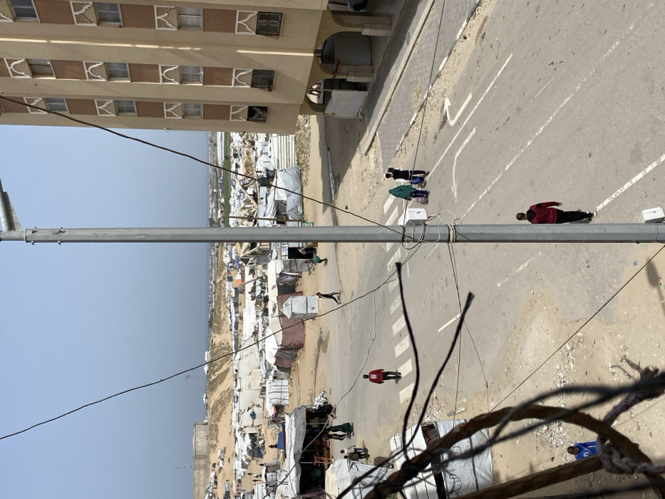 An elevated, outdoor photograph captures a scene in Khan Yunis, Palestinian Territory, depicting a sprawling informal settlement adjacent to a paved road and a multi-story building under a clear, light blue sky.
In the foreground, a prominent gray metallic utility pole extends vertically near the center, with multiple dark electrical wires strung horizontally and diagonally across the lower left portion of the frame, partially obscuring the view. Below the pole, on the road, a male in a red hooded sweatshirt and black pants walks away from the camera.
The midground features an asphalt road with white dashed lane lines and turn arrows painted on the surface; a pedestrian crossing with white stripes is visible further left. To the right of the road, a tall, light brown building with multiple floors is present, featuring numerous windows with decorative white surrounds. Some lower-level windows on this building appear damaged. On the road, near the right, two individuals walk away from the viewer: one wearing a green head covering and dark clothing pulls a light-colored wheeled bag, accompanied by another person in dark attire carrying a bag. Near the pedestrian crossing, a male in a red long-sleeved shirt and dark pants walks left to right across the road.
To the left of the road and extending into the background, a vast area of sandy terrain is occupied by a dense collection of makeshift shelters, tents, and structures made from various materials, predominantly white, gray, and tan tarpaulins. Numerous individuals are visible moving among these structures.
In the background, a sandy ridge or dune rises, with a large, cylindrical concrete structure visible on its summit to the far left. Beyond this ridge, a body of water, consistent with the sea, appears as a light blue band on the horizon. To the right of the sandy ridge, green agricultural fields are also visible extending into the distance.