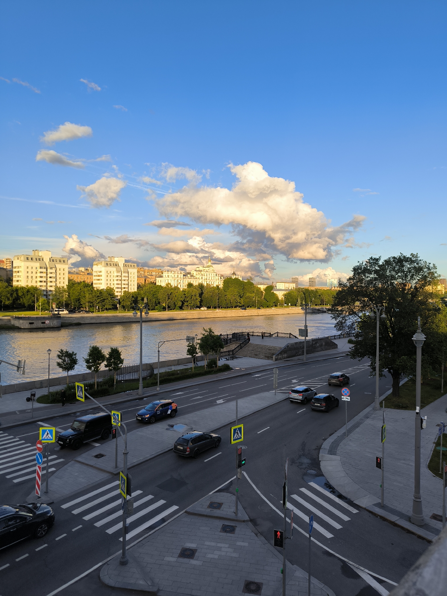 The image captures a city scene at dusk with a wide river in the middle ground, reflecting the golden light of the setting sun and the blue sky dotted with fluffy white clouds. On the far bank of the river, several multi-story buildings and trees form the cityscape. The foreground shows a road with cars and pedestrians. Multiple crosswalks are visible, marked with white stripes on the asphalt. Streetlights and traffic signals are also present. Several cars are driving on the road, and pedestrian crossing signs with yellow squares and a figure walking are prominently displayed. The overall scene suggests an urban environment with active traffic and a touch of natural beauty from the river and sky.
