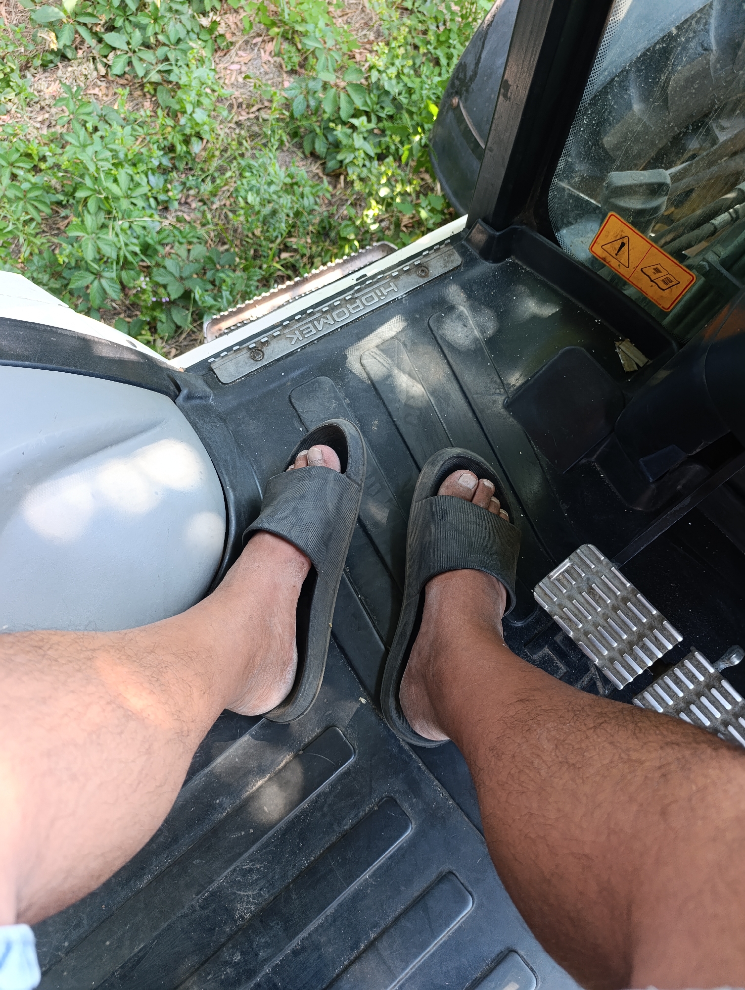 A high-angle, medium shot captures a pair of bare legs and feet wearing black slide sandals, positioned on the floor of a vehicle. The left leg is closer to the camera and appears hairless, while the right leg, further away, is visibly hairy. Both feet are resting on a black, textured floor mat that has "HIDROMEK" embossed on it. To the right, a metal foot pedal with a ribbed surface is visible. The interior of the vehicle is dark and dusty. Outside the vehicle, a patch of lush green foliage and some dry leaves are visible through the open door, suggesting an outdoor or natural setting. The image lacks any specific landmarks to indicate a precise location within Dnipro, Ukraine.