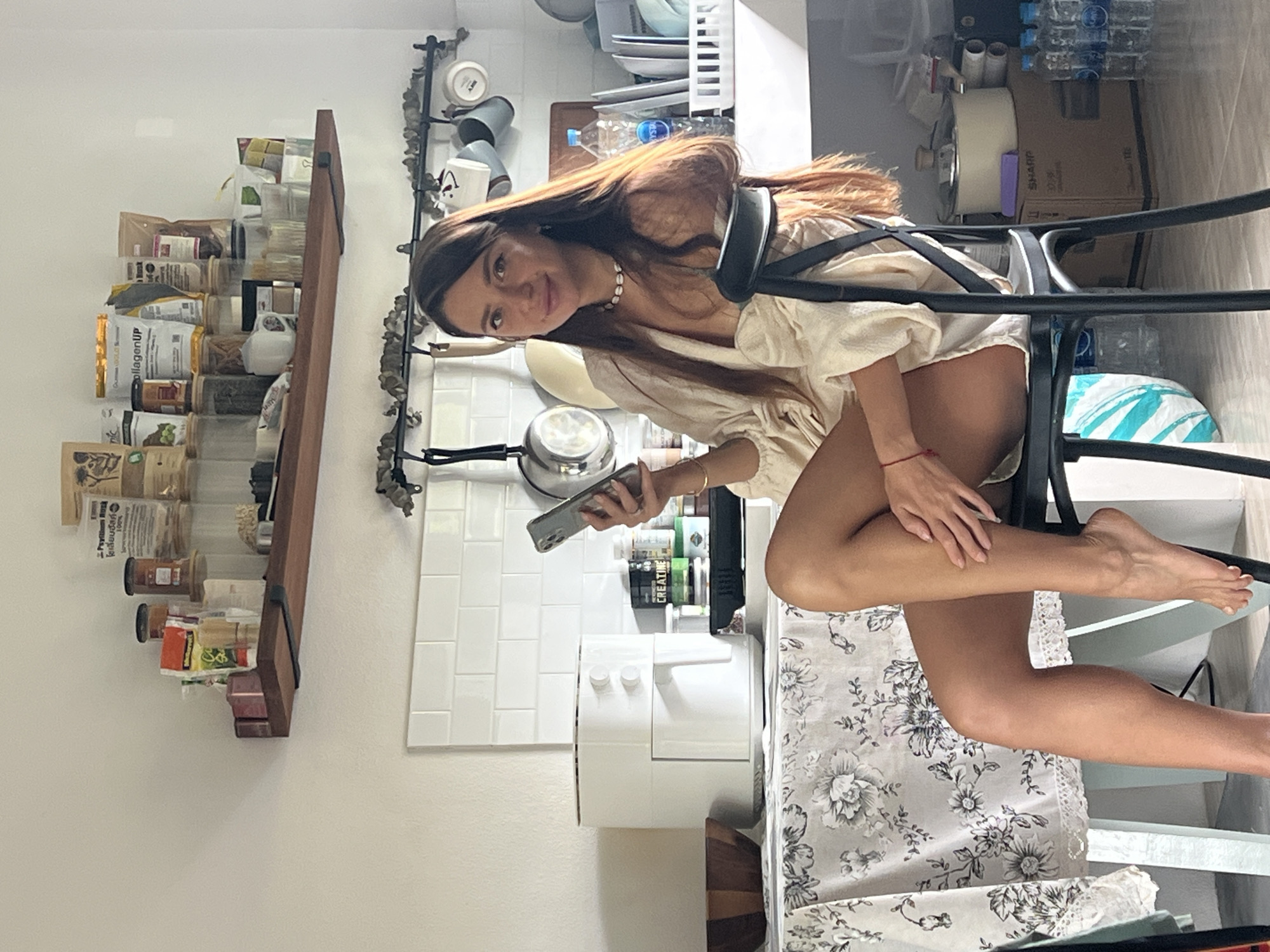 A woman with long brown hair sits on a black chair in a kitchen. She is wearing a light-colored off-the-shoulder top and appears to be holding a cell phone in her right hand. In the background, a white air fryer is visible on a counter, along with various jars and containers on a wooden shelf above. The kitchen has white tiled walls. The scene suggests a home environment in Ban Bang Rak, Thailand.
