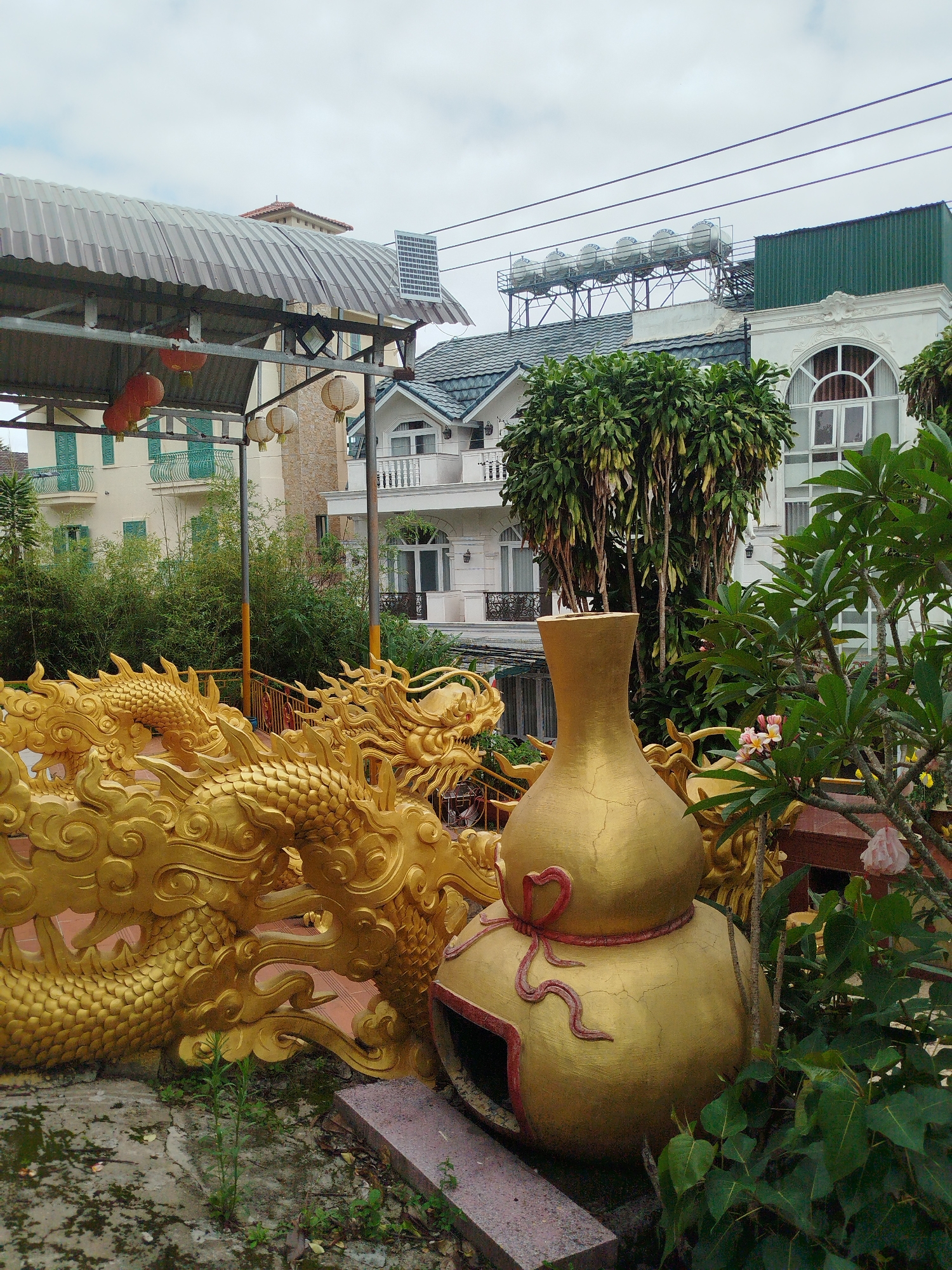 The image captures an outdoor scene in Da Lat, Vietnam, featuring decorative elements and architecture. In the foreground, a large, golden sculpture of a dragon coils on the left, its scales and ornate details illuminated. To the right of the dragon is a golden, gourd-shaped ornament adorned with a red ribbon design, resting on a stone base. Behind these sculptures, a building with a metal, corrugated roof is visible, with red and white lanterns hanging from its structure. Further back, several multi-story buildings with balconies, windows, and tiled roofs dominate the background. Some of these buildings have an Asian architectural influence, with arched windows and decorative moldings. A cluster of trees with lush green foliage stands prominently between the foreground sculptures and the background buildings. The sky is overcast with clouds, suggesting a temperate climate. The ground is paved with concrete and stone, with some patches of overgrown vegetation. The overall scene depicts a decorative area within a city, possibly a temple or a garden, adorned with traditional motifs and modern structures.