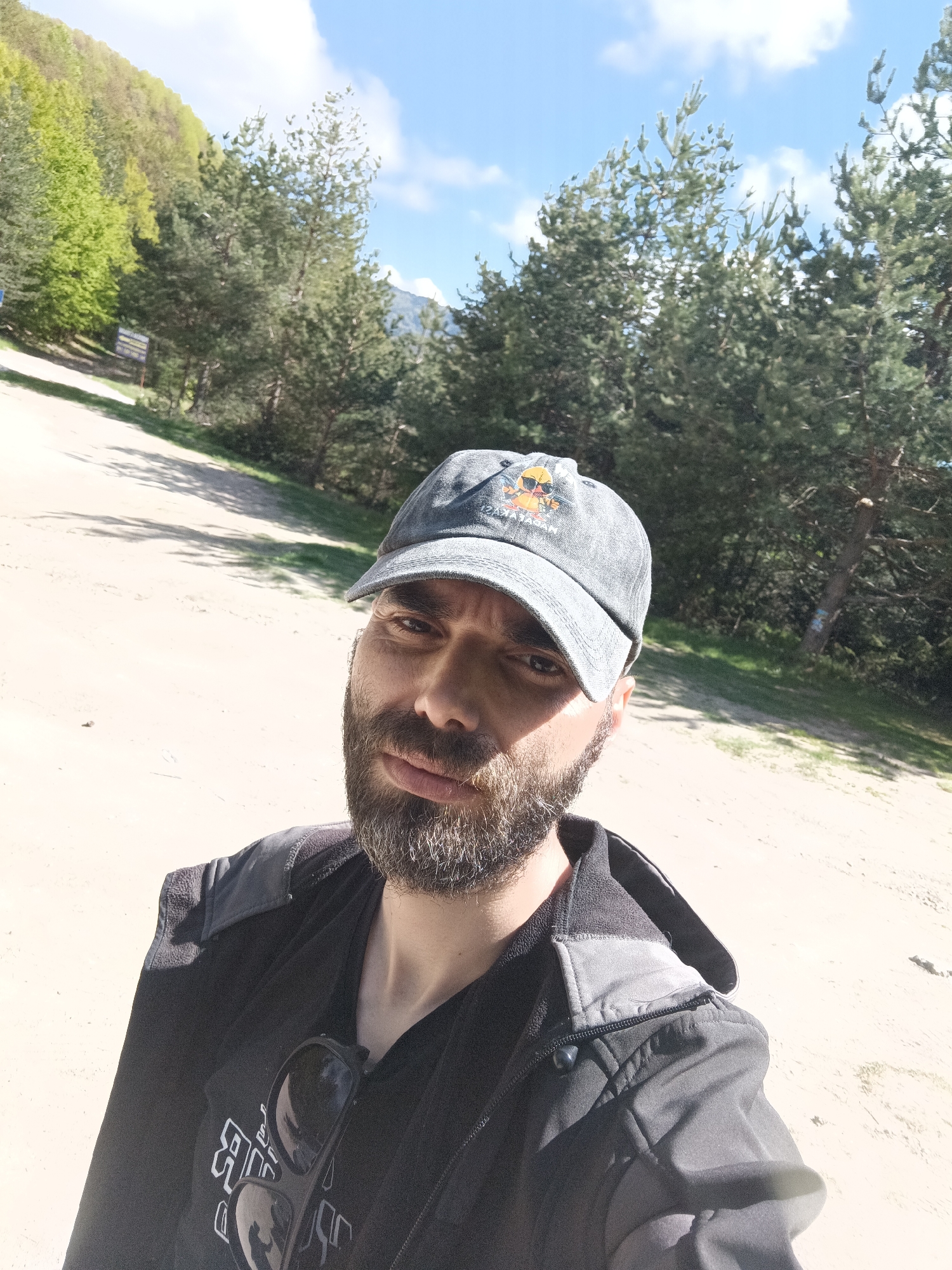 A man with a beard and a cap stands outdoors in Vratsa, Bulgaria. He is wearing a dark jacket over a t-shirt. Sunglasses are hanging from his shirt. The man is standing on a dirt road surrounded by trees and foliage under a partly cloudy sky.