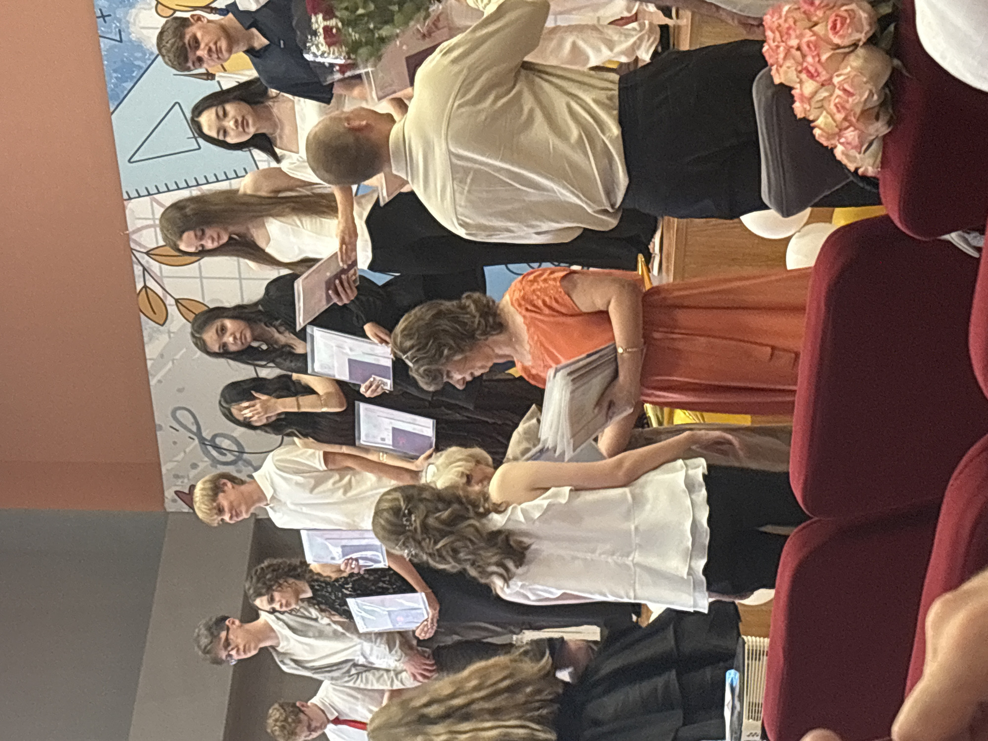 The image captures an awards ceremony on a raised stage decorated with a musical staff motif and geometric shapes. Several individuals, predominantly young women and men, are lined up on the stage, holding certificates or awards.
On the far left, a young man in a gray hoodie and glasses stands next to a young woman in a black lace dress. Next to her is a young man in a white polo shirt. Following him, a young woman with long dark hair wears a black top, and another young woman in a white top and black pants holds a certificate. To her right, a young woman in a white top and black pants is also holding a certificate. Further right, a man in a light-colored shirt and dark pants stands with a bouquet of roses on his right. Another man in a dark polo shirt stands to his right, with a bouquet of red roses in front of him.
In the foreground, a woman in an orange dress stands with a certificate, and to her left, another woman with curly hair in a white ruffled top and black pants holds a certificate. In the lower right corner, a cluster of pink and white roses is visible, and part of a red chair is seen in the foreground, suggesting an audience seating area. Two white balloons and a yellow balloon are also visible near the stage. The lighting appears to be indoor stage lighting.