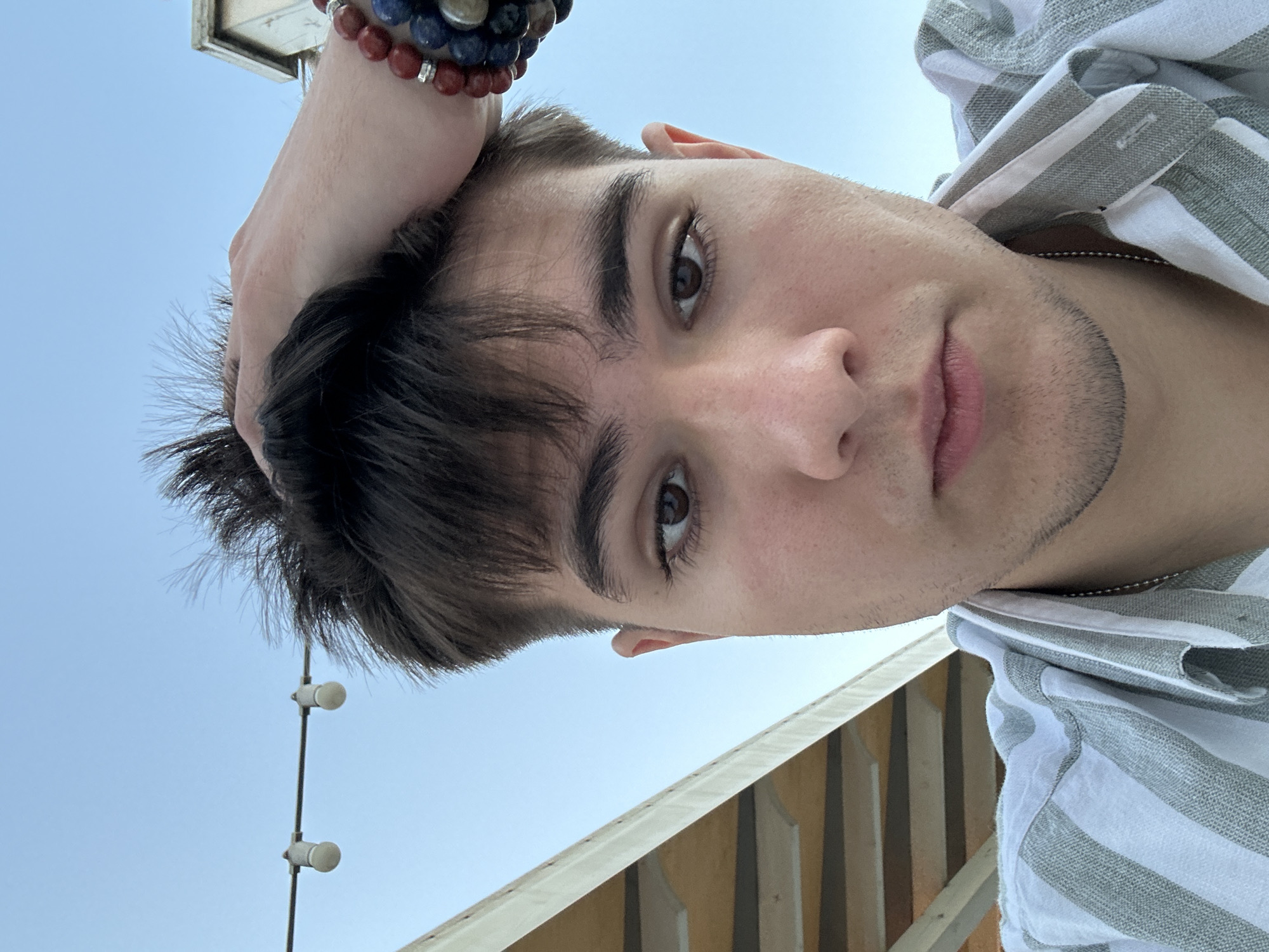 A close-up, low-angle selfie of a young person with dark hair, facing upwards. Their right hand is resting on their head, adorned with a beaded bracelet featuring red, blue, and clear beads. They are wearing a light-colored, horizontally striped shirt with a silver chain visible around their neck. A faint stubble is visible on their chin. The background consists of a clear blue sky and a portion of a building with a light-colored, slatted facade. The lighting suggests it is daytime, likely outdoors. No visible text is present. The image is oriented vertically. The overall impression is casual and personal.