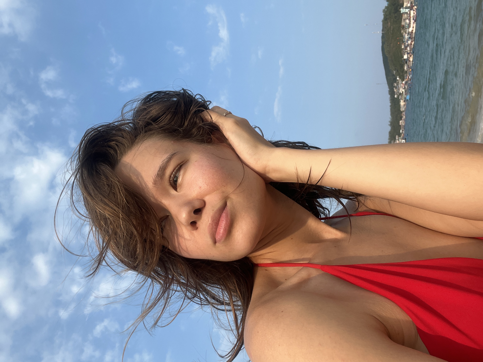 The image captures a young woman taking a selfie outdoors, likely by the sea. She is depicted from the chest up, with her head tilted slightly upwards. She wears a red bikini top and has dark, slightly wet hair that appears wind-swept. Her left hand is raised, gently touching her hair near her ear, revealing a small ring on her finger. Her eyes are partially closed or looking down, and she has a relaxed, subtle smile, conveying a sense of contentment.
The background features a bright blue sky with a few wispy white clouds. To the right, a body of water with small waves is visible, and beyond it, a distant coastline with green hills and scattered buildings. The bright, natural sunlight suggests the time of day is late morning or early afternoon, with clear and sunny weather. The scene clearly indicates a coastal or beach setting, consistent with the provided location context of Nam Natural Heritage Area, Trang, Vietnam. No text is visible in the image.