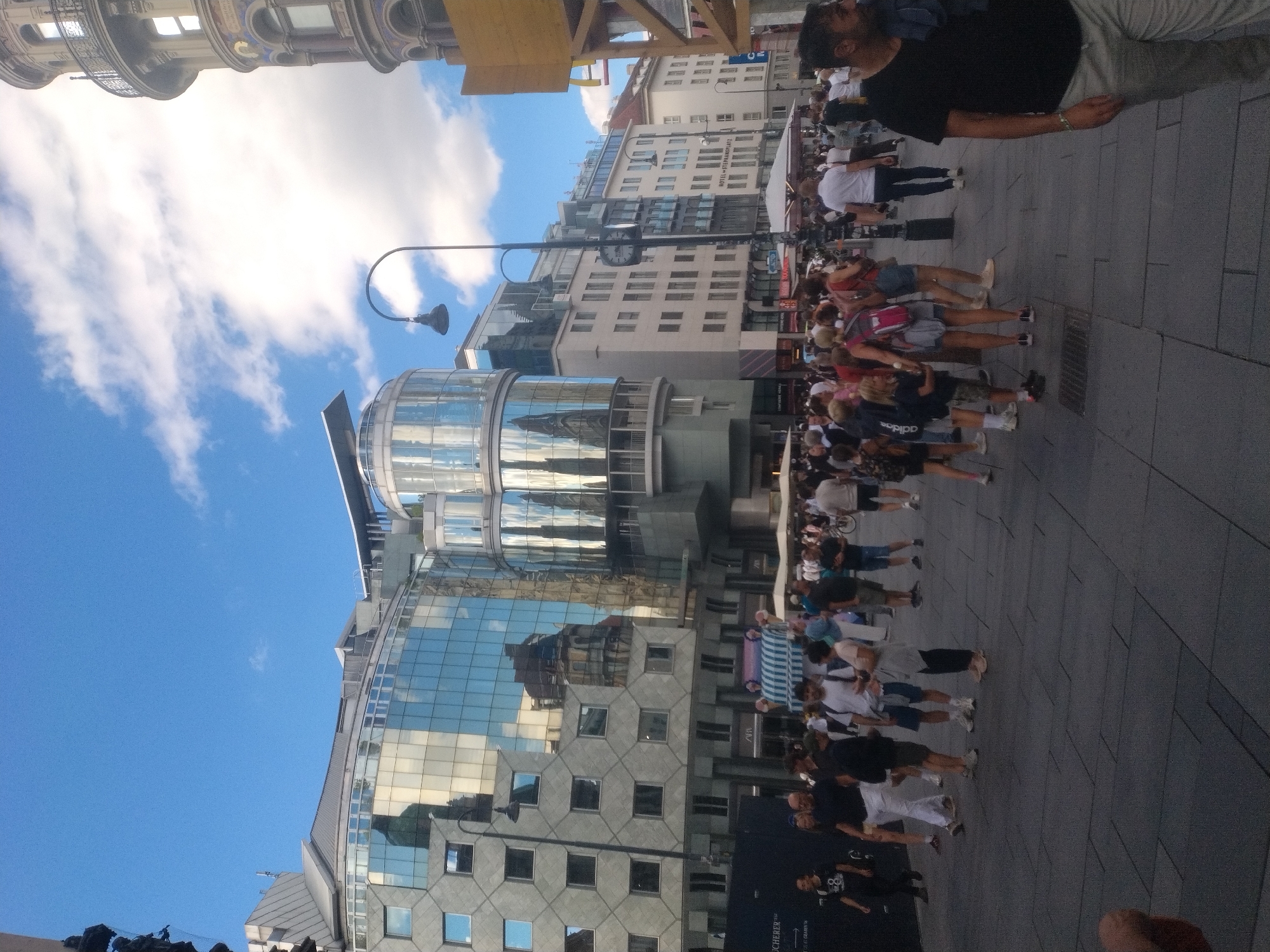 A bright, outdoor daytime scene depicts a street in Vienna, Austria, filled with pedestrians. The architecture features modern glass-fronted buildings with reflective surfaces, juxtaposed with older, ornate buildings and a prominent clock tower. People are walking, standing, and interacting casually. Some are looking at their phones, while others are engaged in conversation. There are signs for shops and businesses visible, including "HOTEL STEPHANPLATZ". The ground is paved with large stone tiles. The sky is blue with scattered white clouds.