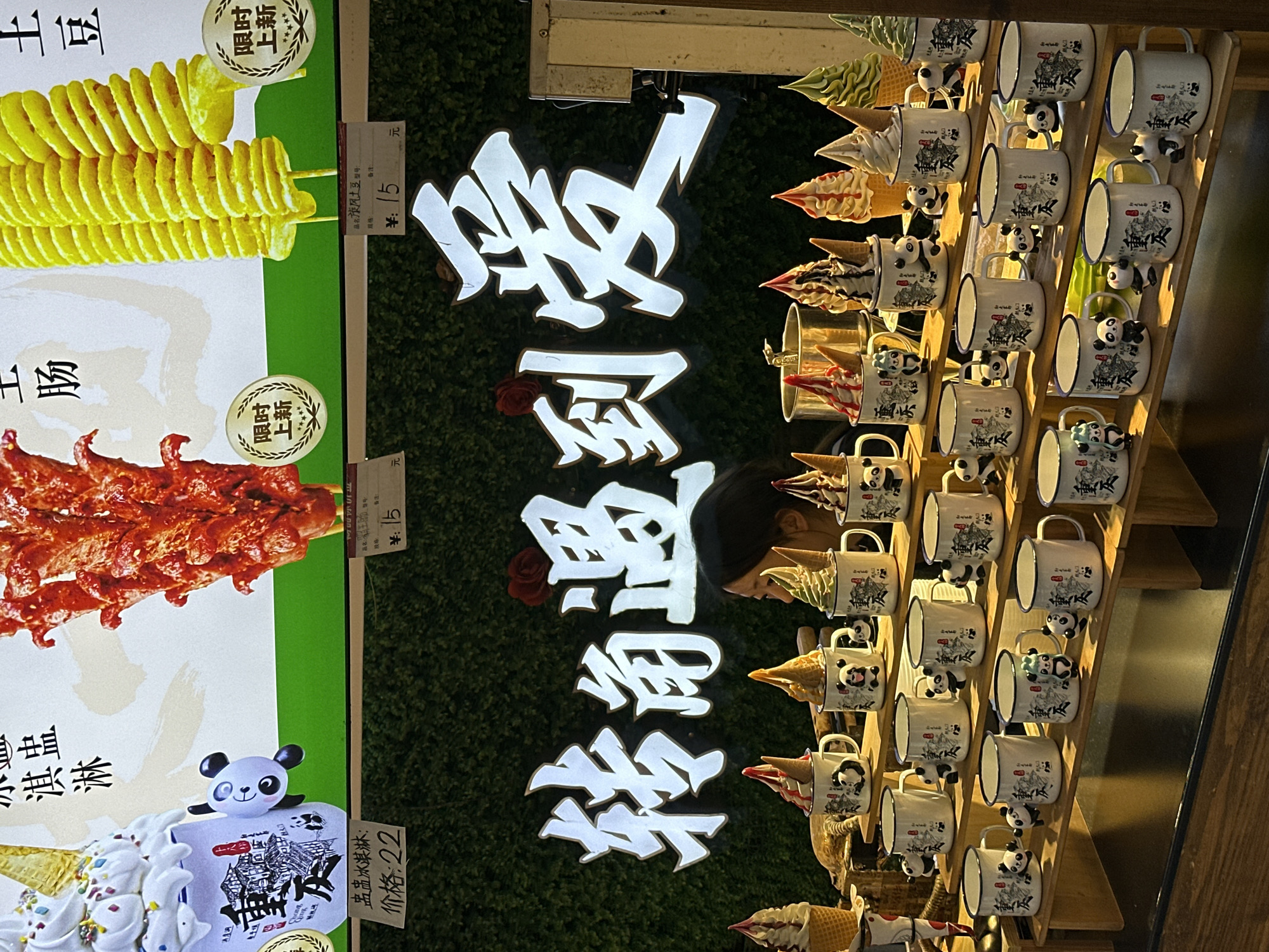 Here is a detailed description of the image:
**Scene:** The image captures a commercial scene at a shop or food stall, possibly located in the Yuzhong District of Chongqing, China. This is indicated by the Chinese characters on the cups in the image.
**Subjects and Objects:**
* **Food Display:** There are images of food items displayed on the left side, likely advertising the stall's offerings. The food items include:
* Spiral-cut potatoes on a stick
* Skewered spicy meat
* Ice cream
* **Pricing:** Pricing information is visible for some of the food items (15 yuan and 22 yuan).
* **Signage:** A glowing sign in Chinese characters that says "转角遇到爱", which translates to "Turning the corner to find love," is displayed.
* **Cups with Ice Cream:** A rack holds white mugs with a blue rim and Panda-themed decorations. Inside are ice cream cones, creating an attractive display.
* **Person:** The back of a person's head is visible as they look at the cup display.
**Actions and Interactions:**
* **Advertisement:** The image seems to be showcasing the various offerings of the stall and potentially attracting customers.
* **Browsing:** The person is observing the mugs and ice cream, likely contemplating a purchase.
**Environment:**
* **Artificial Greenery:** A backdrop of artificial green foliage is used behind the signage, contributing to the stall's visual appeal.
* **Wooden Rack:** The mugs with ice cream are organized on a wooden rack.
* **Lighting:** The scene is illuminated with warm, artificial lighting, particularly highlighting the sign and the ice cream display.
**Overall Impression:** The image shows a vibrant and attractive food stall in what appears to be a commercial area, using appealing displays and signage to attract customers. The Panda-themed mugs are a distinctive feature of the stall.
