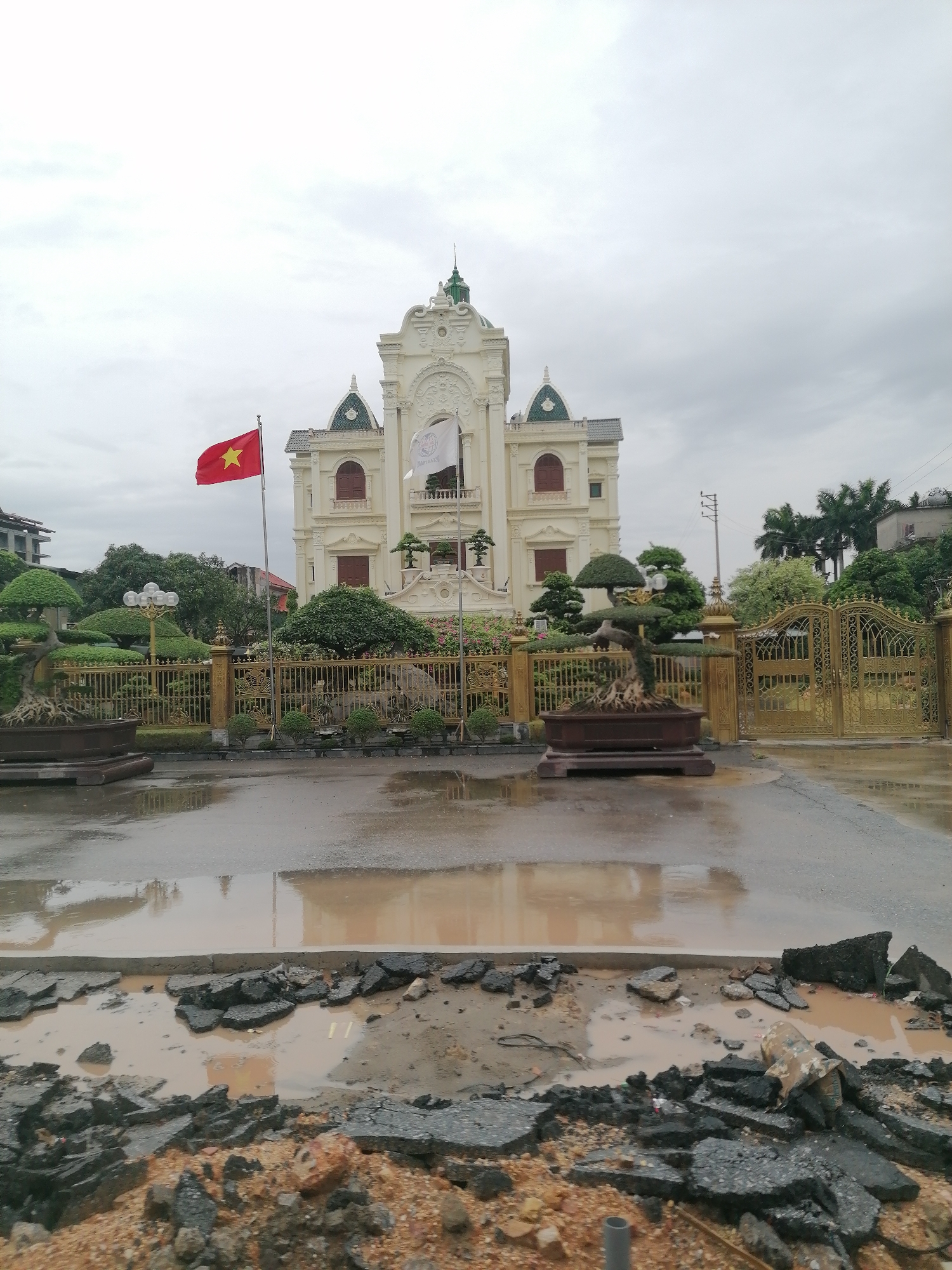 The image depicts a large, ornate white building with a tower in the city of Unknown, Unknown. A Vietnamese flag is prominently displayed on a pole to the left of the building. There is a gold fence surrounding the property. The foreground shows a damaged road with broken asphalt and standing water. The sky is overcast.