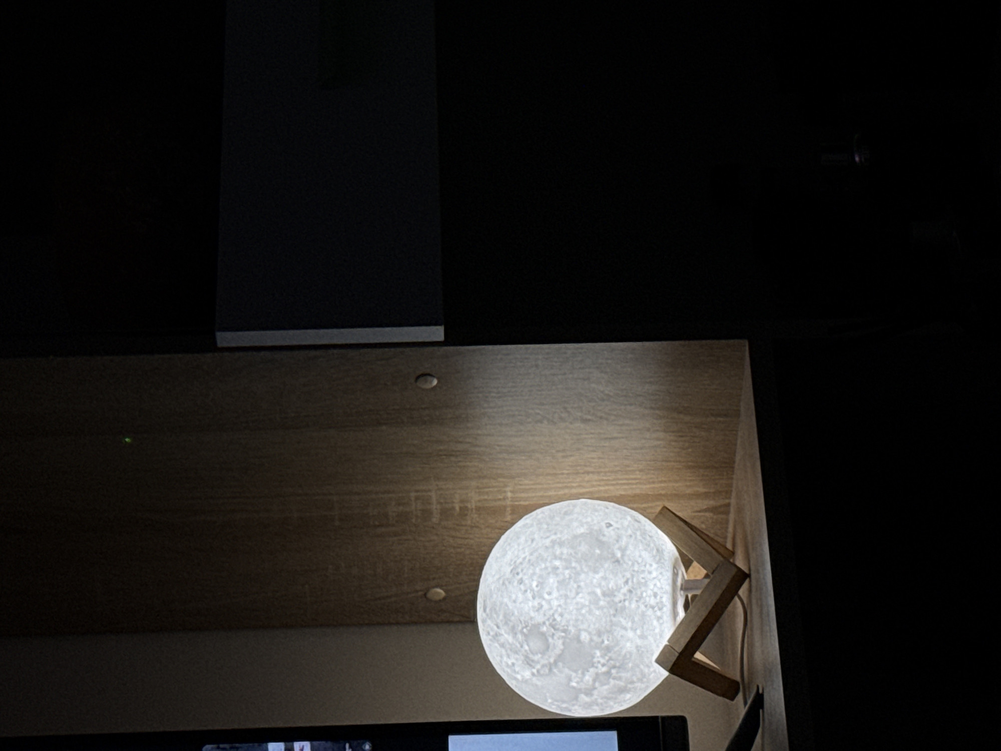 The image presents a close-up view of a room corner with a moon-shaped lamp illuminating the scene. The lamp, a spherical object with a textured surface resembling the moon's craters, is positioned on a wooden stand. The light from the lamp reflects onto the wooden surface above it, creating a warm glow. Above the wooden surface is a dark vertical object, contrasting with the illuminated area. The background is largely dark, suggesting a dimly lit environment.