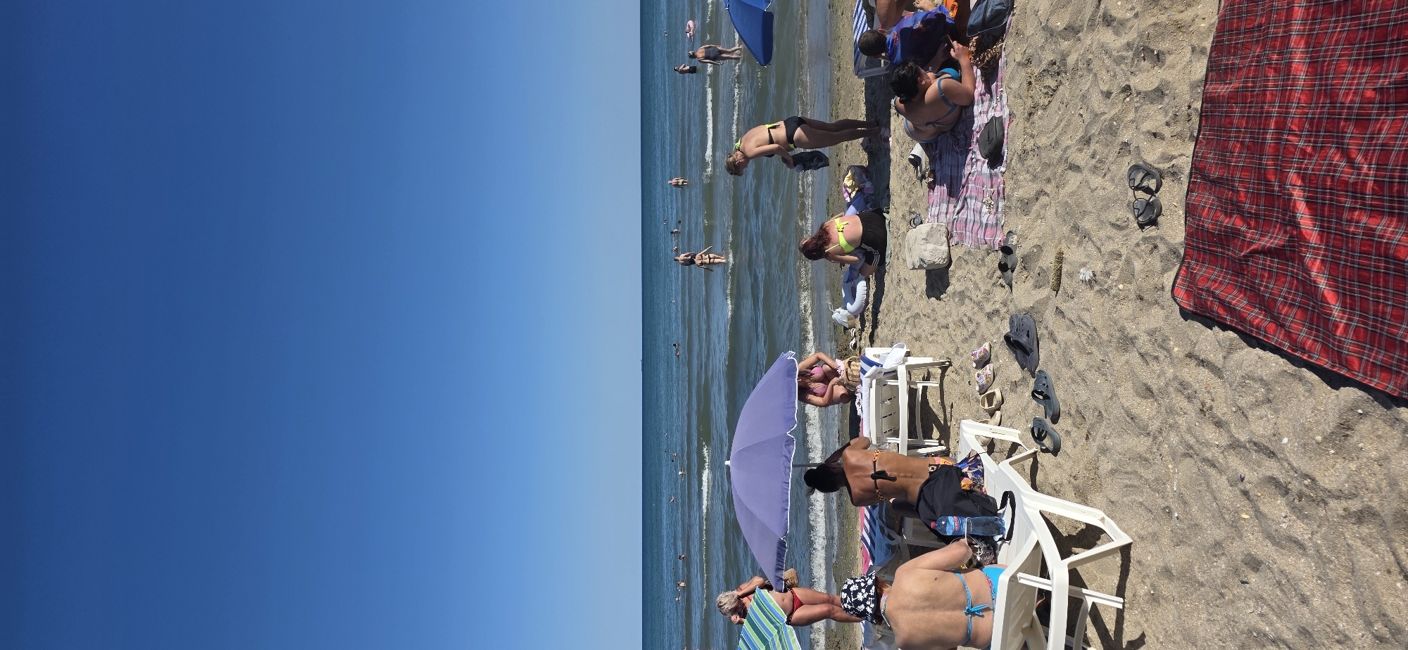 An eye-level, vertical, medium shot captures a busy beach scene with numerous people enjoying a sunny day. The clear blue sky dominates the upper portion of the frame, meeting the calm, blue sea at the horizon line. In the water, various individuals are seen swimming and wading.
On the sandy beach in the foreground, several beach chairs are set up. A purple umbrella provides shade for two people sitting on a lounge chair. To the left, a woman in a red bikini stands next to a green and blue striped umbrella, holding a straw bag. In the center, a woman wearing a black bikini sits on a white lounge chair, facing away from the camera. To the right, a woman in a yellow bikini and black shorts is seated, while another woman in a black bikini stands and appears to be adjusting her clothing. Another person is seated on a patterned blanket. Several pairs of sandals and flip-flops are scattered on the sand, along with a red and black plaid blanket spread out on the lower right side of the frame. A white inflatable ring is also visible.
The sand is light brown and textured, with some scattered debris. The overall atmosphere is one of leisure and recreation on a warm day.