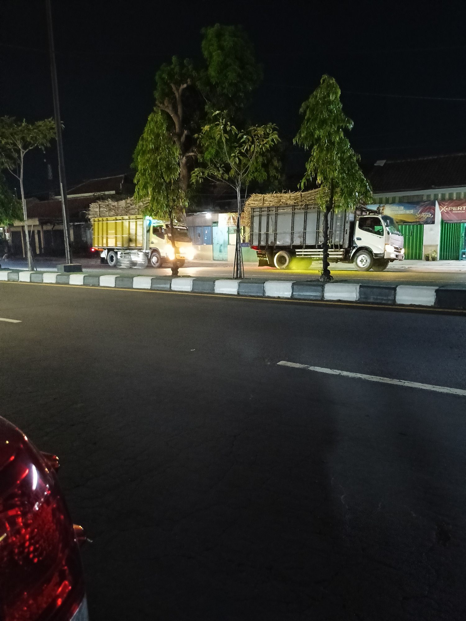 In the city of Kebondalem Kidul, Indonesia, at night, a two-axle white truck carrying a load of sugar cane stalks is parked on a street. The truck is facing to the right. Its headlights are on, illuminating the road and the truck itself. To the left of this truck, another yellow truck with its headlights on and carrying a similar load of sugar cane stalks is also parked on the street, facing to the right. The street is paved with asphalt and bordered by a curb with white and gray markings. In the background, there are buildings, trees, and a dark night sky. A portion of a red vehicle's taillight is visible in the lower left corner of the frame.