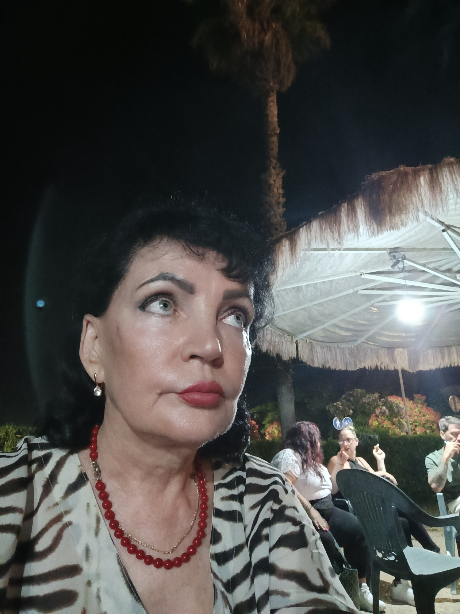 The image shows a woman in the foreground, looking up and to the right. She has dark hair, light-colored eyes, and is wearing red lipstick and a red beaded necklace. Her clothing has a black and white striped pattern. In the background, other people are seated at tables, and a large umbrella with fringe is visible. The scene appears to be outdoors at night, possibly a restaurant or cafe, with a palm tree in the background. There is no specific indication of the city of Giulianova, Italy, within the image itself.