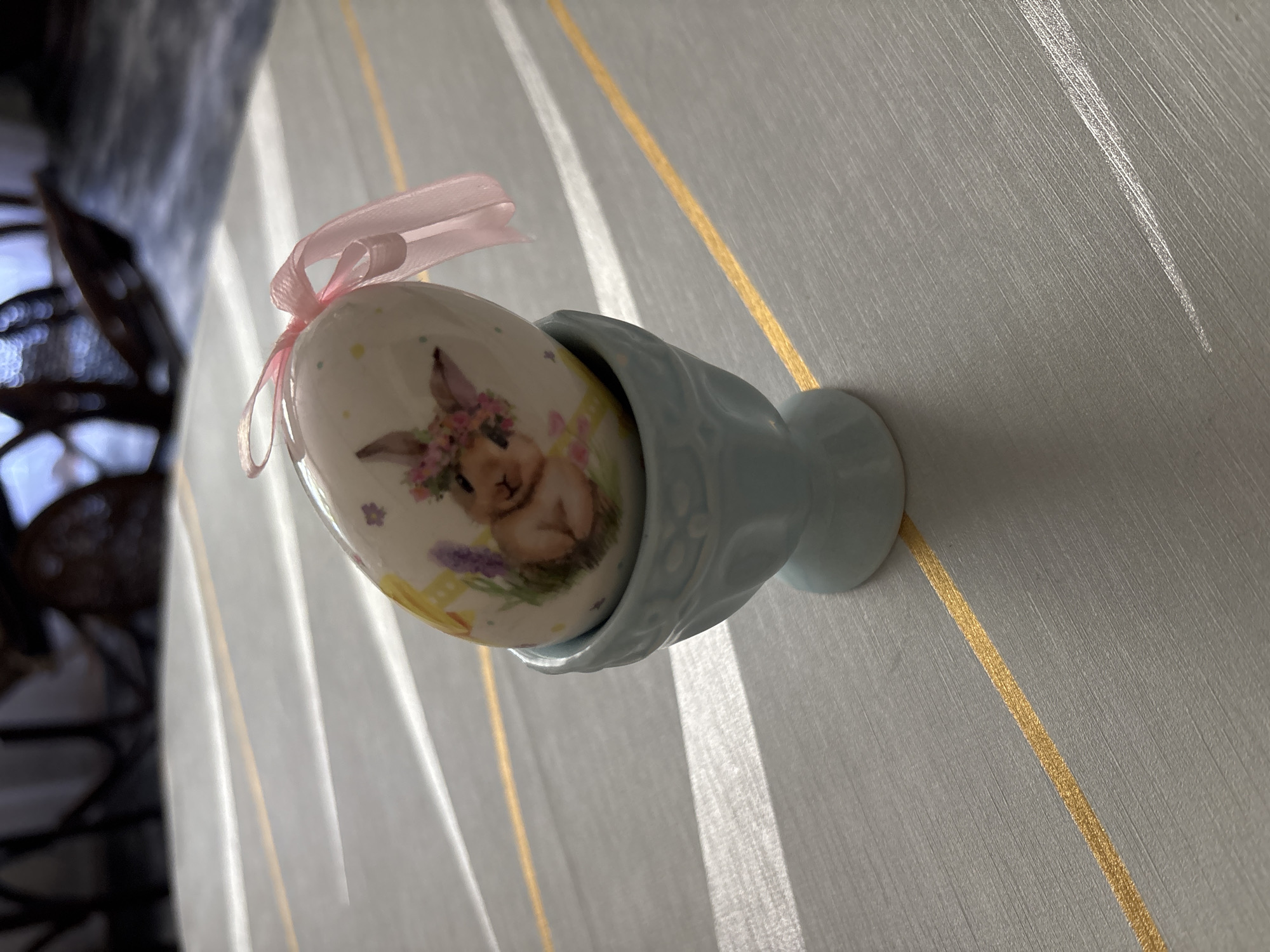 The image displays a single decorative Easter egg, positioned in a light blue ceramic egg cup, resting on a light grey, textured surface marked with thin, horizontal gold stripes. The egg is white, featuring an illustration of a light brown rabbit adorned with a floral wreath on its head, accompanied by small lavender flowers and yellow accents. A pink satin ribbon is tied into a bow at the apex of the egg. Scattered around the main design on the egg are small yellow, light blue, and pink dots. Distinct parallel bands of strong light and shadow fall across the grey surface, originating from an unseen source to the upper left. The background is softly blurred, showing dark, indistinct shapes that suggest indoor furniture. This scene is situated in the city of Chernogorsk, Russia.