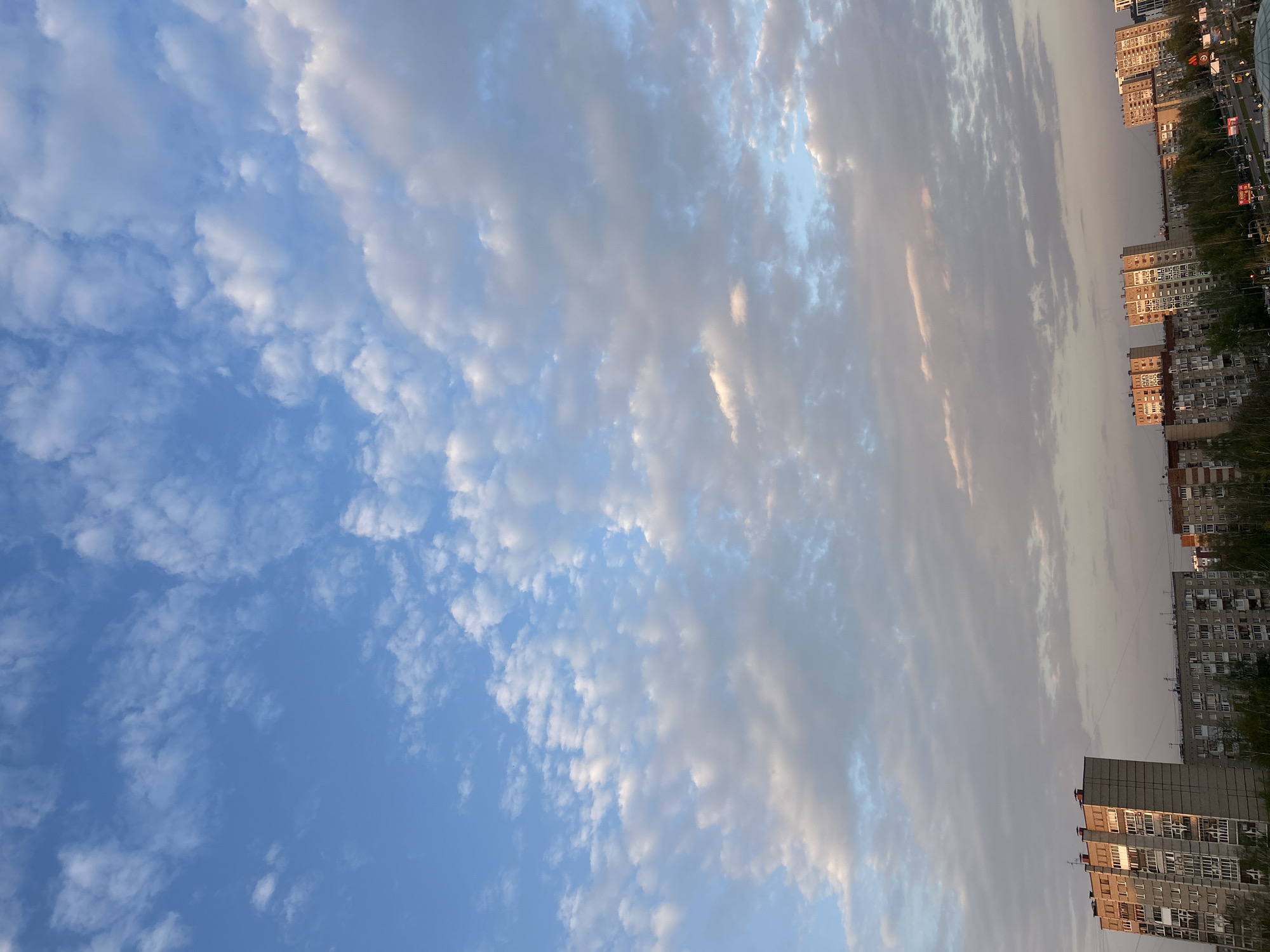 The image shows a view from Izhevsk, Russia, oriented with the sky to the left and the cityscape to the right. The sky is predominantly blue with scattered patches of small, white cumulus clouds. To the right, the clouds transition to a more uniform gray layer, possibly indicating an approaching weather change. Below the clouds, several tall apartment buildings are visible. The buildings are typical of Soviet-era architecture, featuring many windows and a rectangular shape. Trees are planted along the street, and a few vehicles are present, suggesting a residential area within the city.