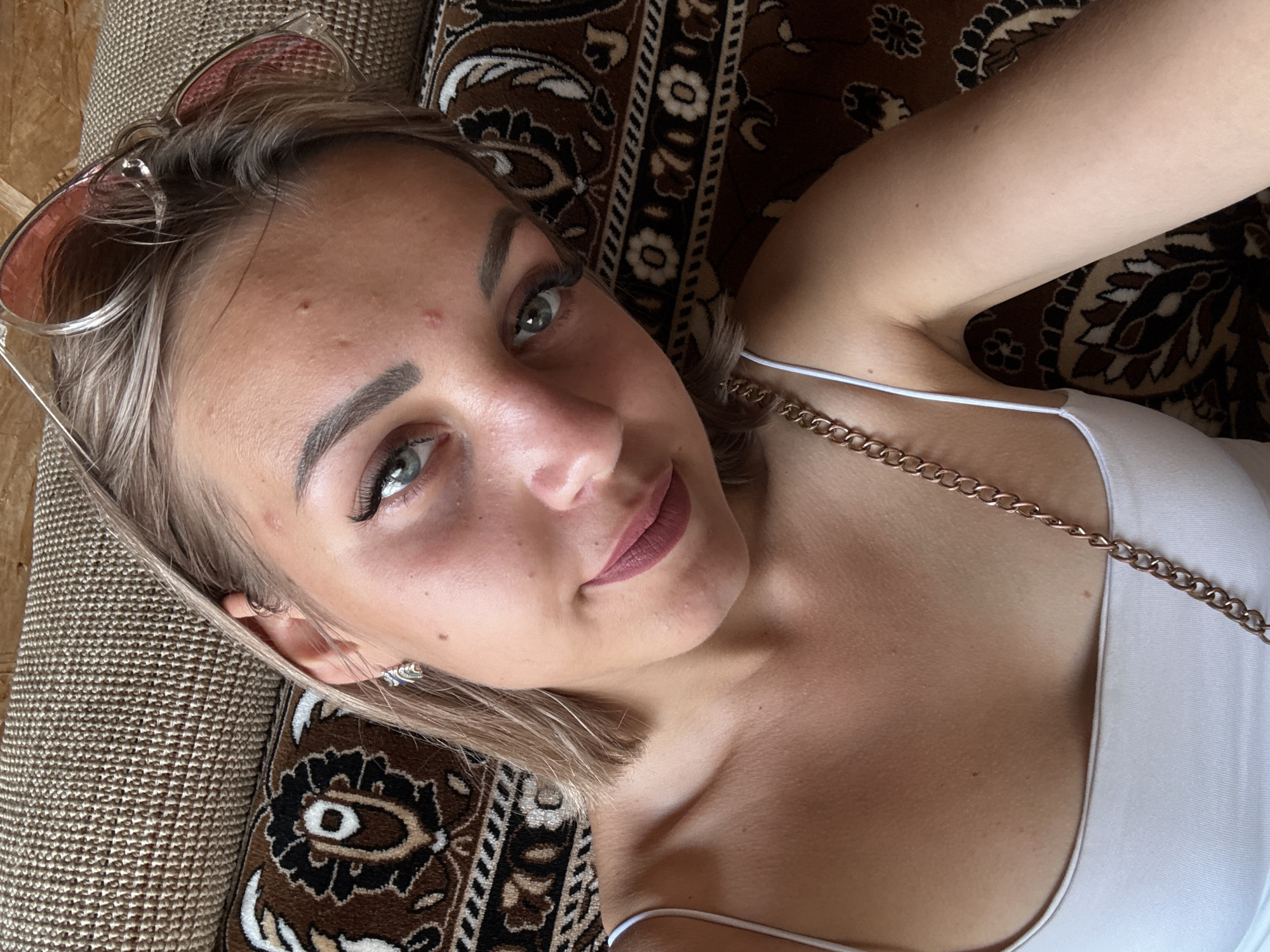 A young woman with blonde hair and green eyes is lying on a textured brown sofa. She is wearing a white tank top and a gold chain necklace. Her sunglasses are perched on her head. The sofa has a patterned fabric with brown and beige colors. The image is a close-up selfie. The woman appears to be in a domestic setting, possibly her home. The city of Krasnogorskoye, Russia is not visible in this image.