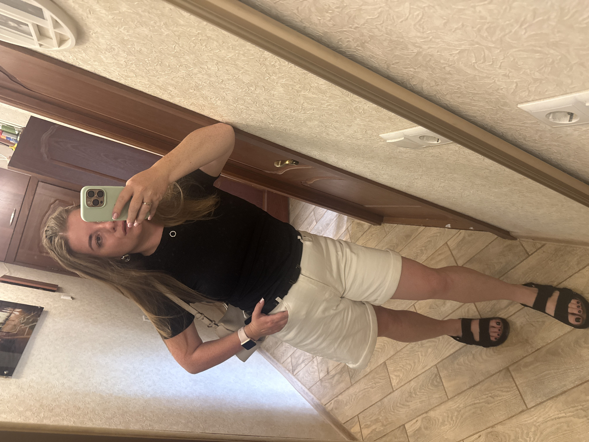 A woman with long, wavy blonde hair stands in what appears to be a hallway, taking a mirror selfie with her phone. She is wearing a black t-shirt, white shorts, and black sandals. A light-colored purse is slung over her left shoulder. Her right hand is in her pocket, and her left arm is resting against a wooden doorframe. The hallway has light-colored wood-paneled flooring and textured walls. A framed picture is visible on the wall to her left, and a tall wooden cabinet is to her right. The lighting is moderate, casting some shadows.