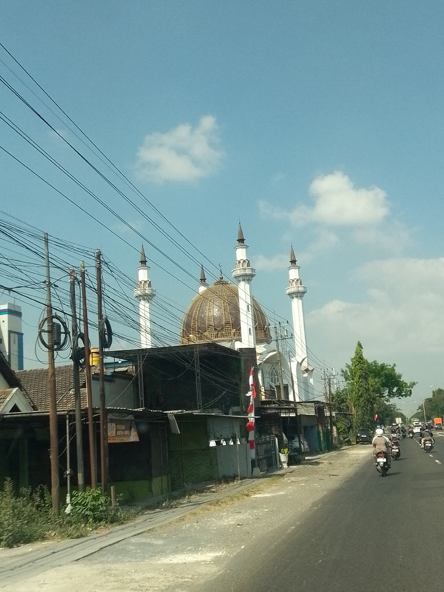 The image captures a street scene in Plosowahyu, Indonesia, featuring a prominent mosque with multiple minarets and a large golden dome. The mosque is located on the right side of the street, with its distinctive architecture clearly visible. In the foreground, a road runs parallel to the mosque, with several motorcycles and their riders traveling in the same direction. Utility poles and a dense network of overhead wires are visible in front of the mosque and along the side of the road, indicating urban infrastructure. The sky is blue with scattered white clouds. Buildings and vegetation are present along the street, contributing to the urban landscape.
