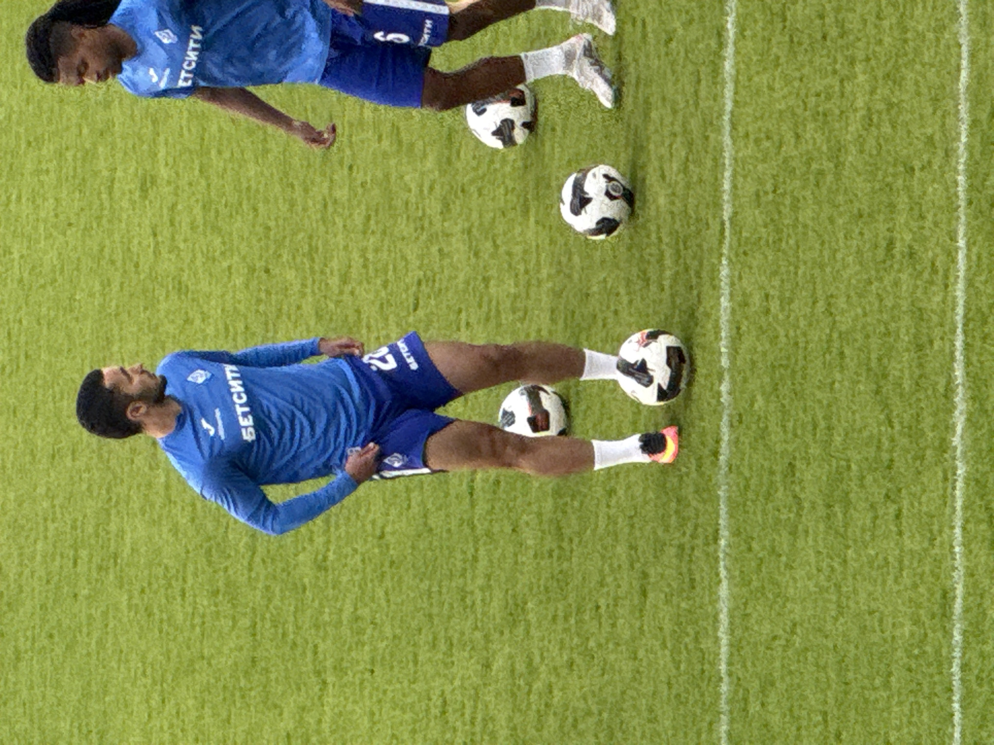 The image shows two soccer players on a green field in what appears to be a practice session. The field has white lines marking boundaries or zones. Four soccer balls are scattered on the field. The players are wearing blue uniforms and white socks. One player is positioned at the top of the frame, while the other is lower in the frame. The word "БЕТСИТИ" is visible on the shirts of both players. The location could be Yekaterinburg, Russia, due to the Cyrillic lettering on the shirts, which translates to "BETCITY."