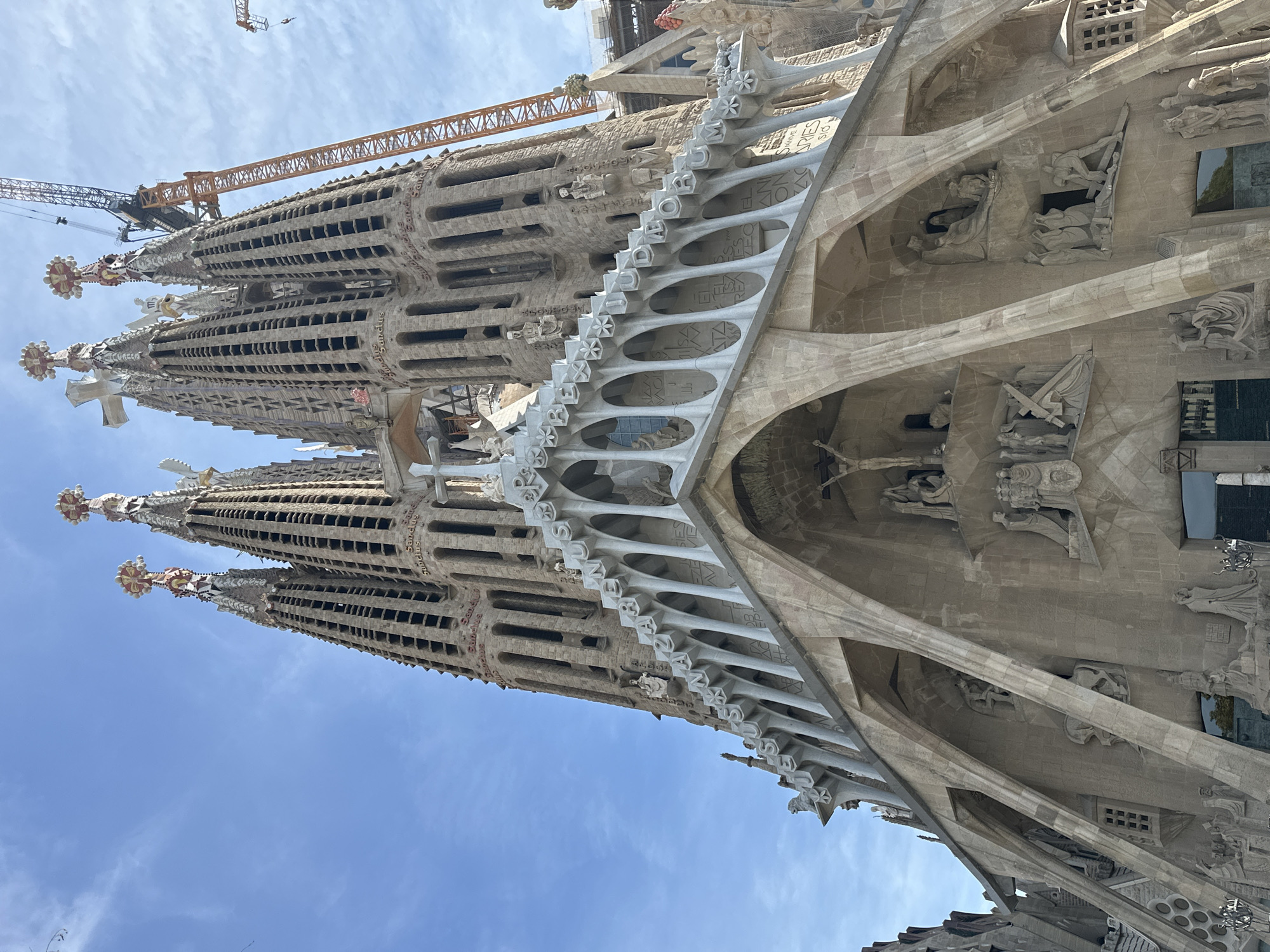 This image depicts the Passion Façade of the Sagrada Família basilica, located in Barcelona, Spain. The photograph is taken from a low-angle perspective, emphasizing the height and intricate details of the structure against a partially cloudy blue sky.
The central subject is a massive stone basilica, characterized by its highly detailed and angular architecture. The lower section of the façade features three prominent arched entrances. Above these entrances, numerous sculptures in a cubist-inspired style depict scenes from the Passion of Christ. A large crucifix with the figure of Christ is centrally positioned above the main entrance. Other sculptural groups portray figures carrying a cross, lamentation scenes, and other related narratives.
Above the main sculptural area, a horizontal architectural element forms a balustrade or architrave, featuring repeating star-like or floral motifs and an inscription that reads "JESUS NAZARENUS REX IUDAEORUM" (Jesus of Nazareth, King of the Jews). This element supports higher sections of the façade and the bases of the bell towers.
Rising prominently are several tall, tapering bell towers, constructed from light-colored stone. These spires are perforated with numerous vertical openings and bear repetitive inscriptions of the Latin word "Sanctus" (Holy). The apex of each visible spire is crowned with ornate, multi-colored finials, some appearing cross-shaped.
To the right and rear of the basilica, multiple large construction cranes are visible, primarily orange and black in color. One particularly tall lattice-boom crane extends high above the existing spires, indicating ongoing construction activities. The background sky is a clear blue with scattered thin, white cirrus clouds. The scene is illuminated by daylight, creating subtle shadows that enhance the three-dimensional relief of the architecture and sculptures.