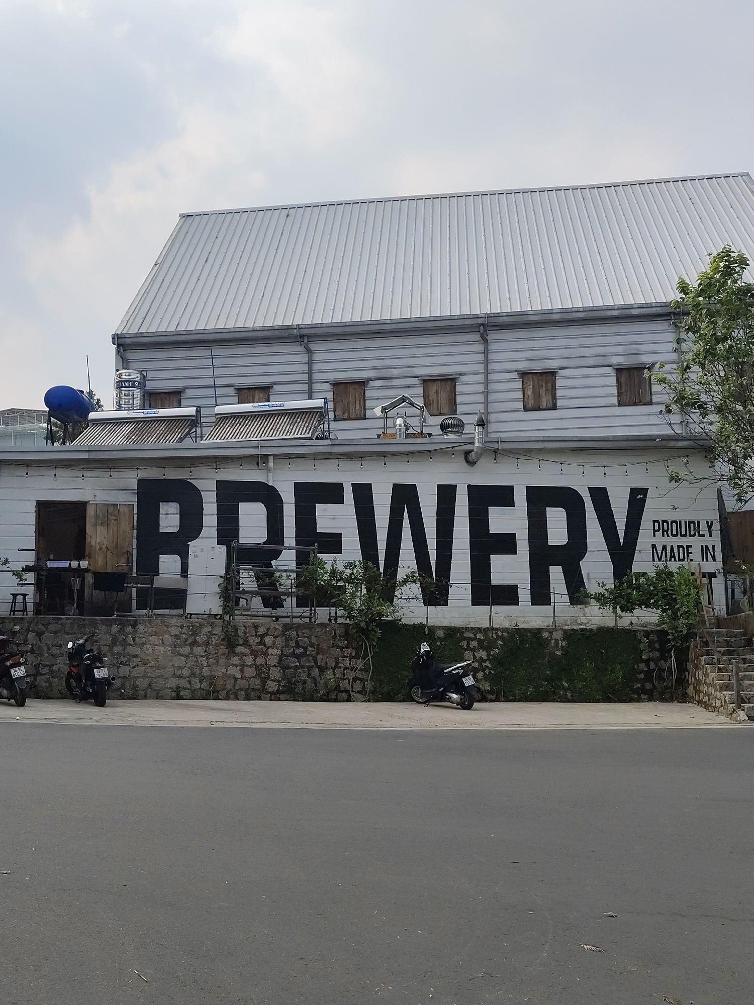A daytime exterior shot shows a building with the word "BREWERY" painted in large black letters on its white facade. Below it, in smaller letters, is "PROUDLY MADE IN". The building has a corrugated metal roof and small, shuttered windows on the upper level. A stone wall runs along the base of the building, with greenery growing on it. Several scooters are parked along the curb in front of the building. The building is located on a street in Da Lat, Vietnam.