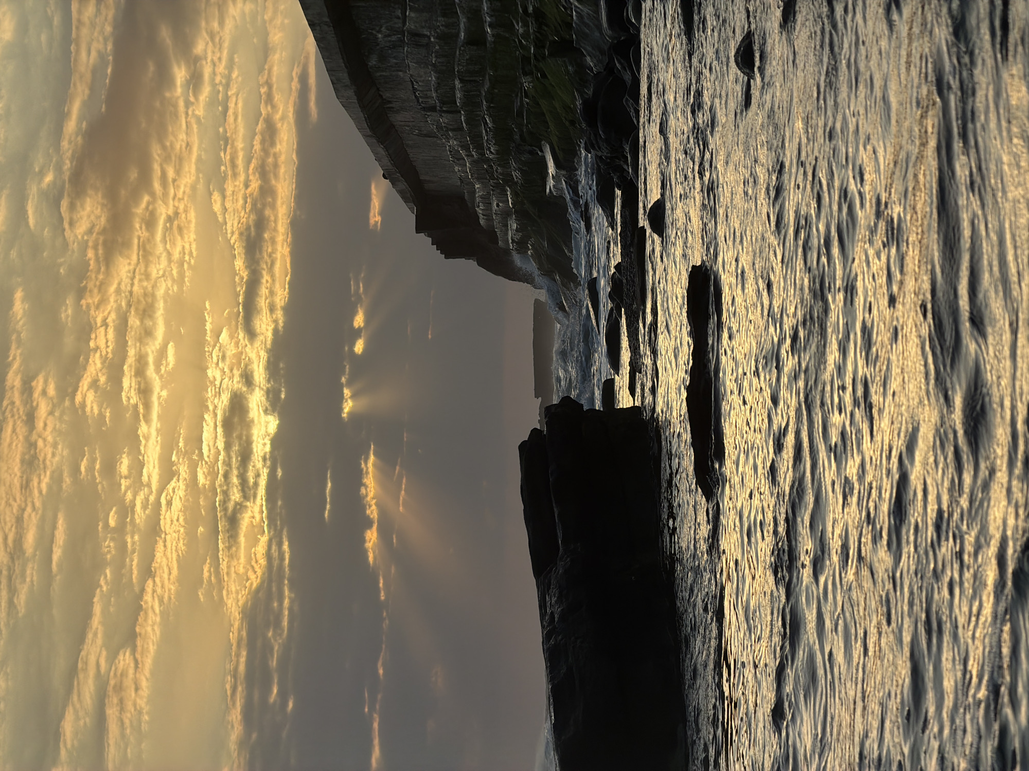 A rocky coastline meets a concrete seawall under a cloudy, golden-hour sky. The sun breaks through the clouds, casting dramatic rays onto the water and the seawall. Waves crash against the rocks and the base of the wall. The water is choppy and reflects the warm light. The overall scene is one of natural beauty and man-made construction interacting with the elements.