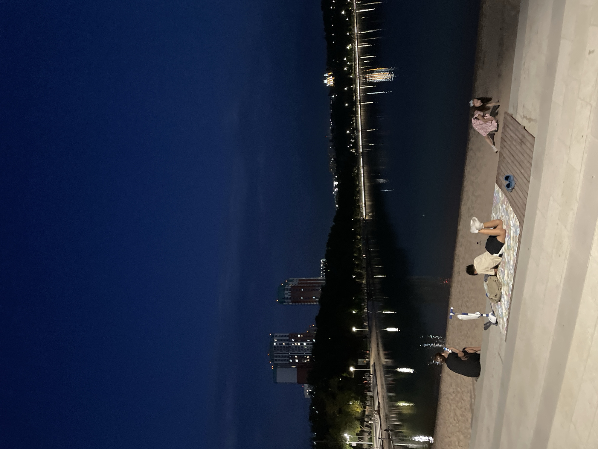 In Almaty, Kazakhstan, at dusk, a serene scene unfolds by a body of water. The sky is a deep, dusky blue, transitioning to black as night approaches. In the foreground, a sandy embankment meets a paved walkway with tiered steps. A person in a black shirt and shorts sits on the sand, facing away from the camera, looking towards the water. A white scooter stands nearby. Further along the sand, two individuals are relaxing on a mat. One person, wearing a light-colored shirt and dark shorts, lies on their stomach, looking towards the water. Another person, in a pink dress, is on their hands and knees on the sand. Across the water, the cityscape is illuminated by lights from buildings and streetlights lining a tree-lined promenade. Reflections of these lights shimmer on the dark water, creating a picturesque urban landscape.