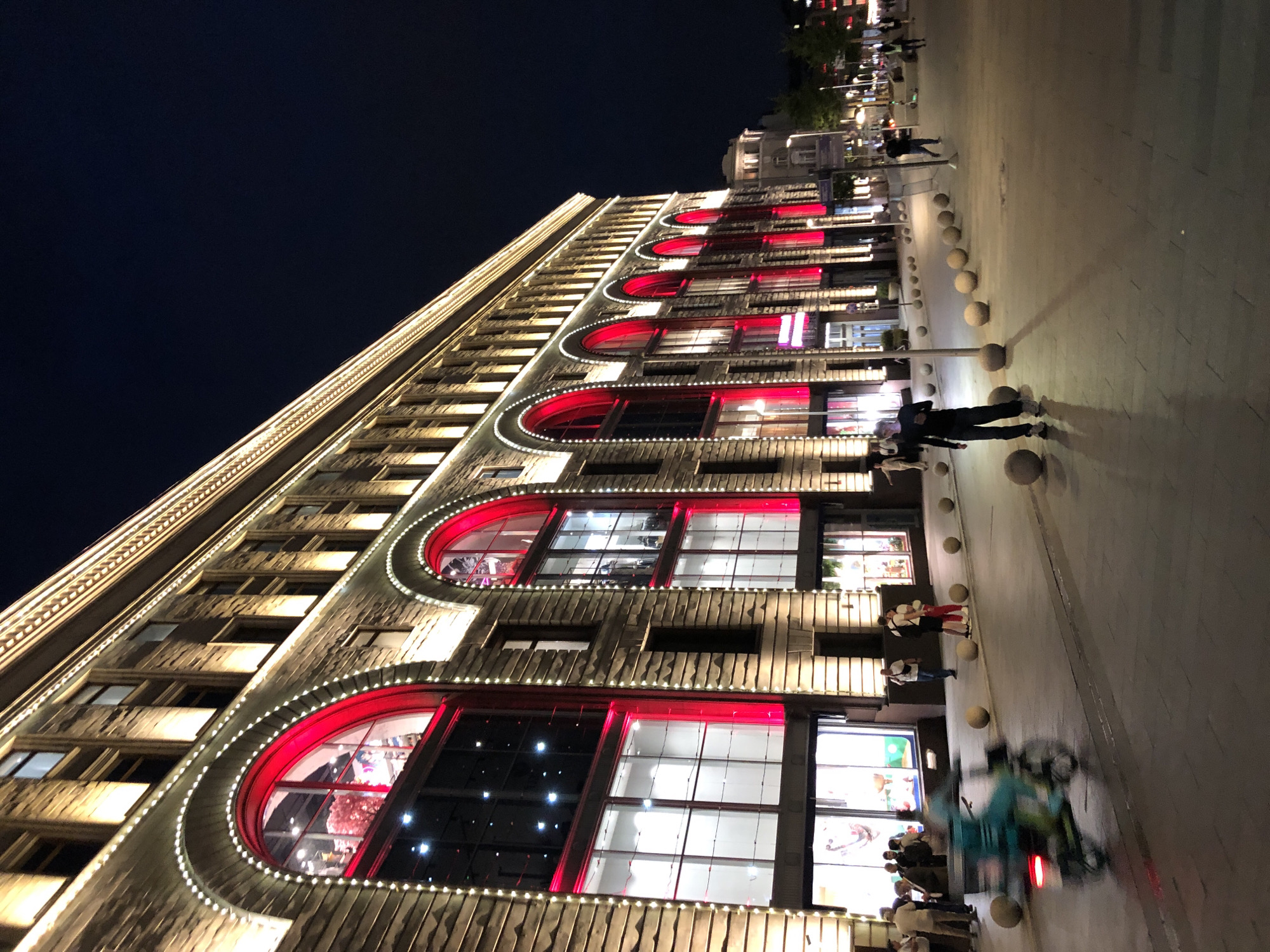 The image depicts a street scene in Moscow, Russia, at night. A large, ornate building with illuminated arched windows featuring vibrant red interiors dominates the left side of the frame. The facade is adorned with string lights. On the sidewalk in front of the building, several people are walking, standing, and a scooter rider is in motion, blurred by movement. The street is paved with dark stones, and a row of spherical bollards lines the sidewalk. In the background, other buildings and streetlights are visible, along with trees and a dark night sky.