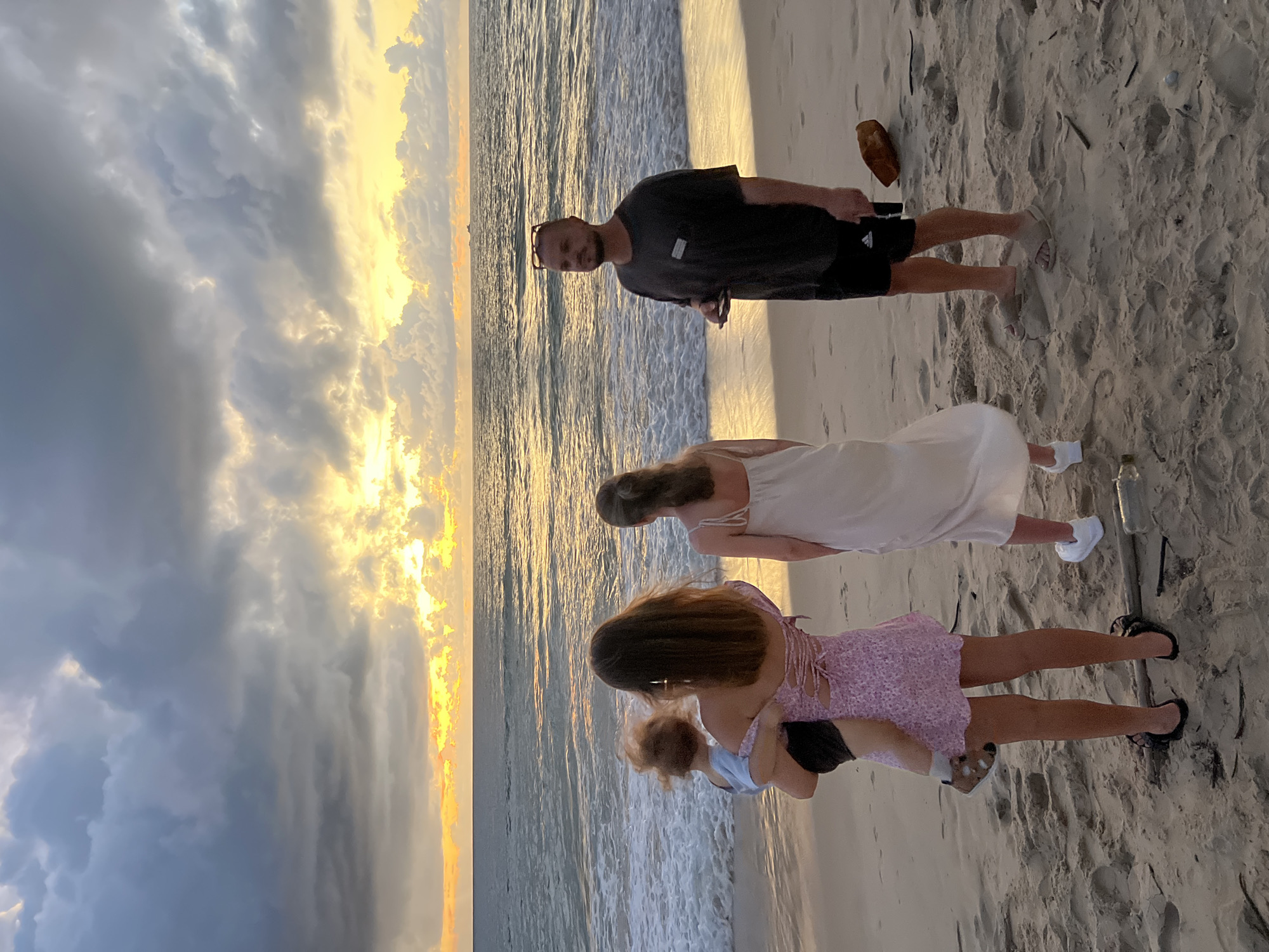The image shows a group of four people on a sandy beach. The group consists of one man and three women or girls. The man is wearing a dark t-shirt and shorts, and is standing on the sand. He is looking towards the camera. The three women or girls are facing away from the camera, towards the ocean. The woman closest to the man is wearing a white dress. The next woman is wearing a pink floral dress. The youngest person is wearing a light blue top. They are standing in shallow water at the edge of the ocean. The sun is setting, casting a warm, golden light on the clouds and the water. The sky is a mixture of dark grey clouds and bright yellow and orange hues, indicating a dramatic sunset. The ocean is calm with gentle waves. The sand is light brown and has some debris, possibly seaweed, scattered on it. The scene appears to be a casual outing on a beach at sunset, possibly in Choeng Thale, Thailand. The overall mood seems relaxed and serene. There are no discernible signs or text visible in the image.