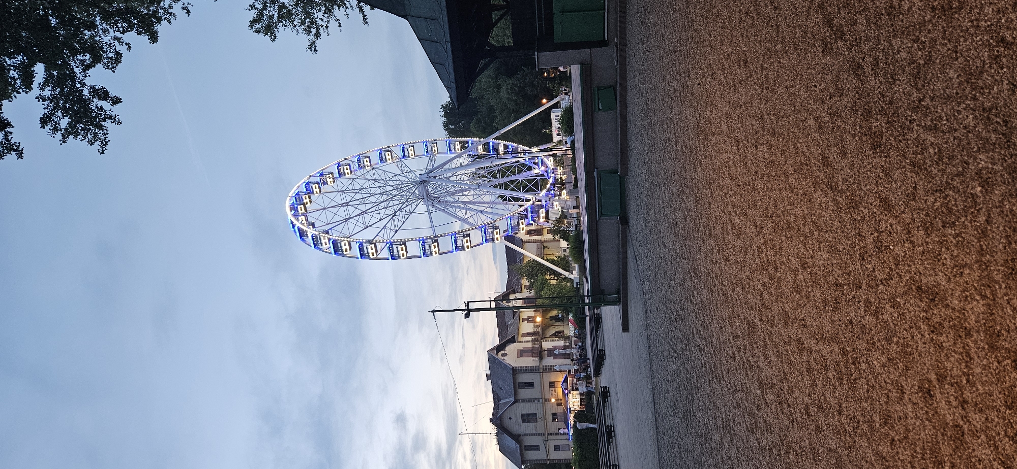 This image depicts a Ferris wheel illuminated with blue and white lights at dusk in Keszthely, Hungary. The Ferris wheel is the primary subject, positioned in the center of the frame, its circular structure reaching towards the twilight sky. Below and to the left of the Ferris wheel, a row of buildings with red-tiled roofs and balconies is visible. These buildings appear to be part of the town's architecture, possibly housing businesses or residences. Scattered in the foreground and midground are various elements of an outdoor public space, including gravel-covered ground, benches, and trees. To the right, a dark, roofed structure, possibly a gazebo or pavilion, is present, partially obscuring the background trees. Several people are subtly visible, some seated at tables outside one of the buildings, suggesting a recreational or dining area. The overall scene conveys a peaceful evening atmosphere in a town with a blend of natural elements and man-made attractions.