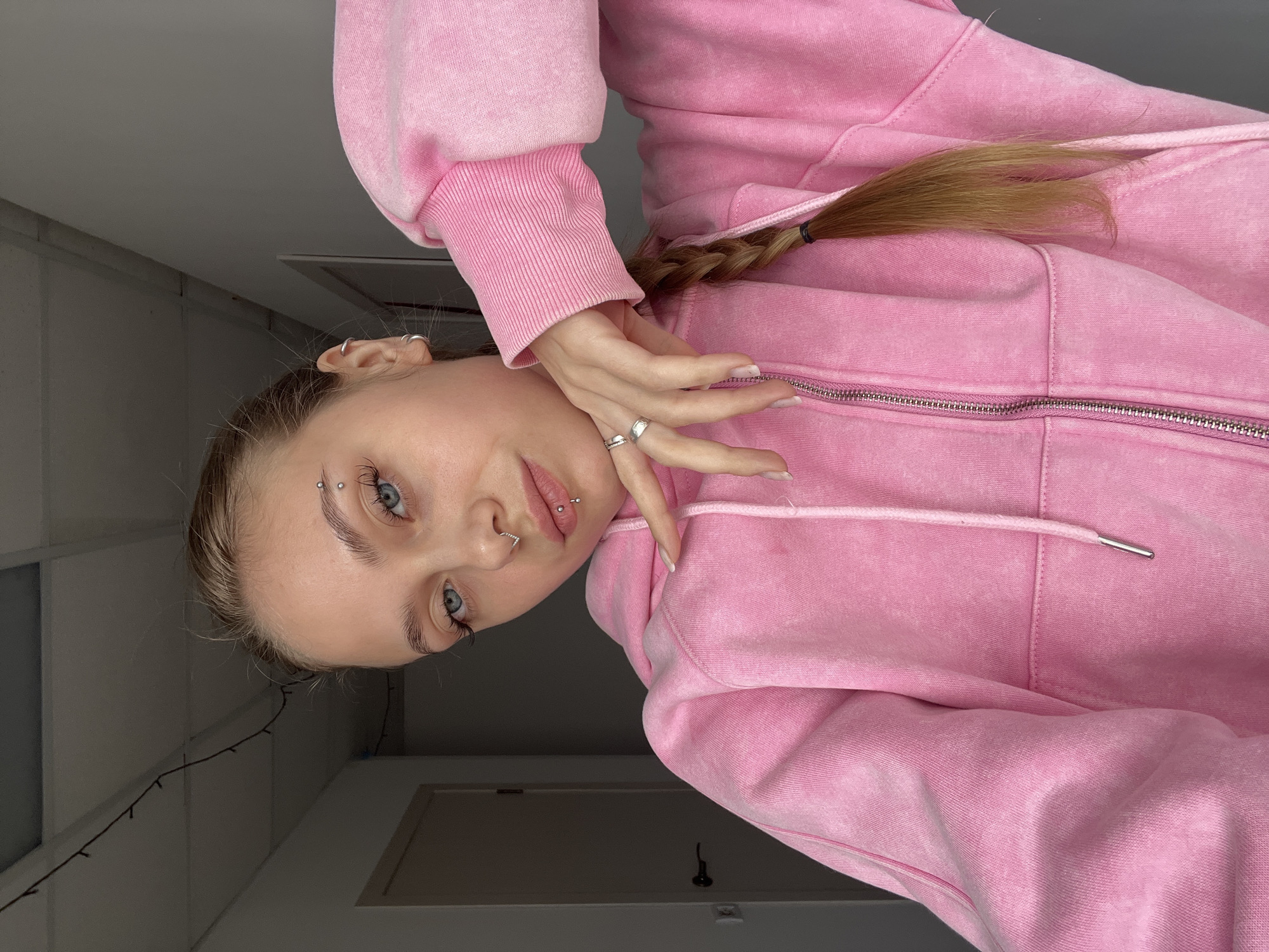 A young woman with blue eyes and pierced eyebrows and lips is facing the camera, wearing a pink zippered hoodie. Her blonde hair is pulled back in a braid. She is holding her hand up to her neck, with a ring on her finger. The background is a light grey wall with a white door. The image is rotated counter-clockwise. The image was not taken in Фоки, Russia.