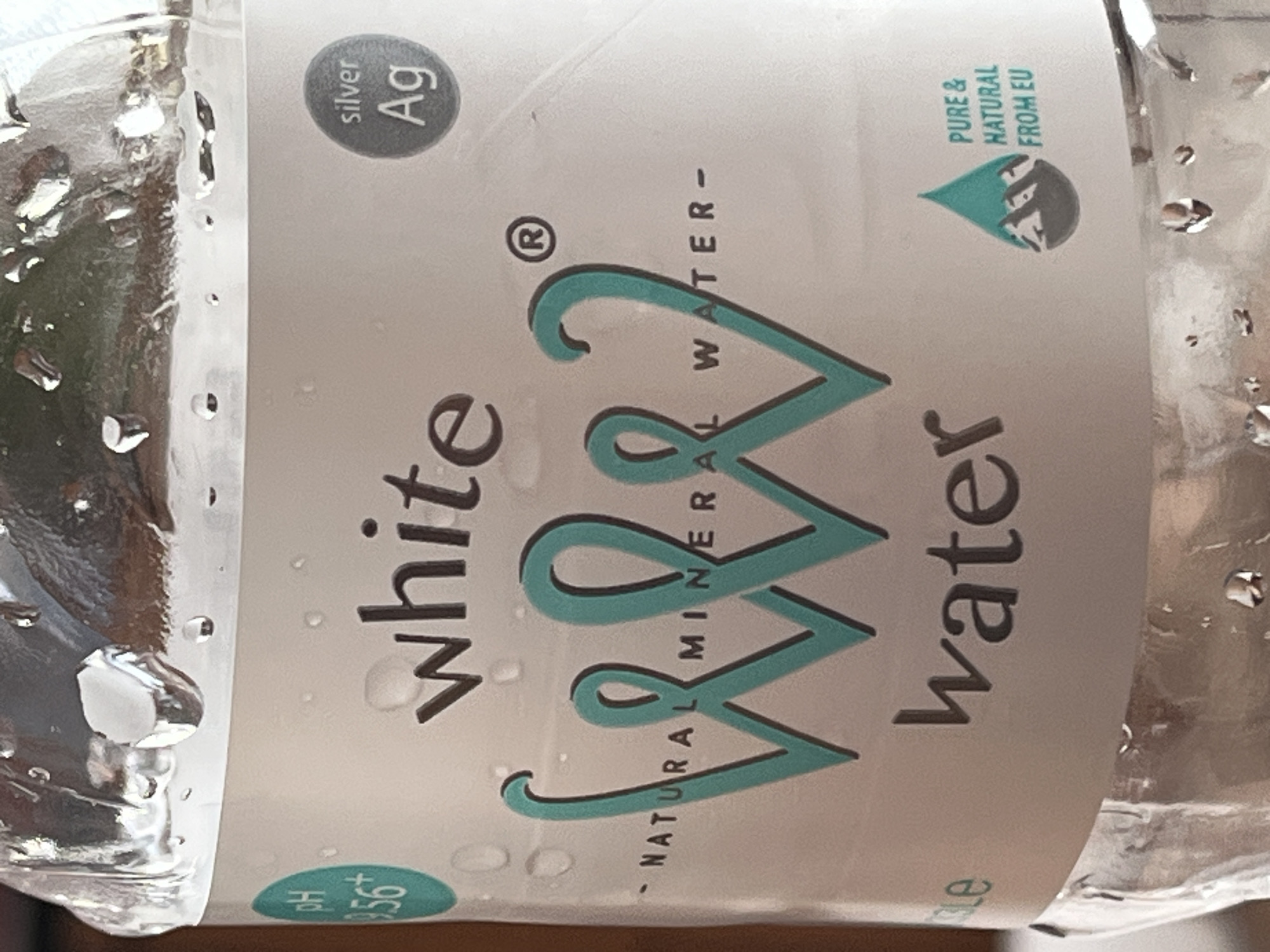 The image shows a close-up of a plastic bottle of White Water, a natural mineral water. The bottle has a white label with the brand name "white" in dark lettering. Below this, a stylized "WWW" logo in a teal color is prominent. Beneath the logo, the words "- NATURAL MINERAL WATER -" are printed in black. To the left of the "WWW" logo, there is a teal circle with "pH 8.56+" inside. To the right of the logo, there is a dark gray circle with "silver Ag" inside. Towards the bottom right of the label, a teal teardrop-shaped icon contains a silhouette of mountains and the text "PURE & NATURAL FROM EU". The bottle is slightly out of focus and has condensation on its surface. The background is blurred and appears to be indoors with warm lighting. The image does not provide any information about the city of Vratsa, Bulgaria.