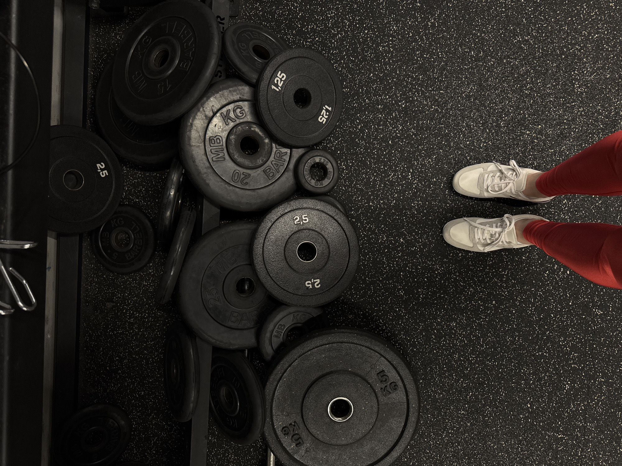 This high-angle, overhead shot captures a person's lower body standing on a speckled black rubber gym floor, surrounded by various barbell weight plates. The perspective looks down at the person's feet and the weights, which are mostly arranged on a weight rack system on the left side of the frame.
On the right, the person wears white sneakers with white laces and bright red athletic leggings. Their feet are positioned on the black and white speckled floor, suggesting they are either pausing during a workout or preparing to begin an exercise. No specific activity is clearly depicted beyond standing.
The left and bottom portions of the image are dominated by numerous black, circular weight plates of varying sizes. These plates are stacked or stored within a metallic rack, portions of which are visible. The plates are clearly marked in kilograms ("KG"), indicating a metric system environment. Visible weight increments include "1.25", "2.5", "5", "10", "15", "20", and "25" kilograms. Many of the plates prominently feature the brand "MB BARBELL". The overall scene depicts an indoor fitness facility or gym setting. Based on the provided context, this scene is located in Sochi, Russia, a detail further supported by the "MB BARBELL" brand, which is associated with Russian fitness equipment manufacturers.