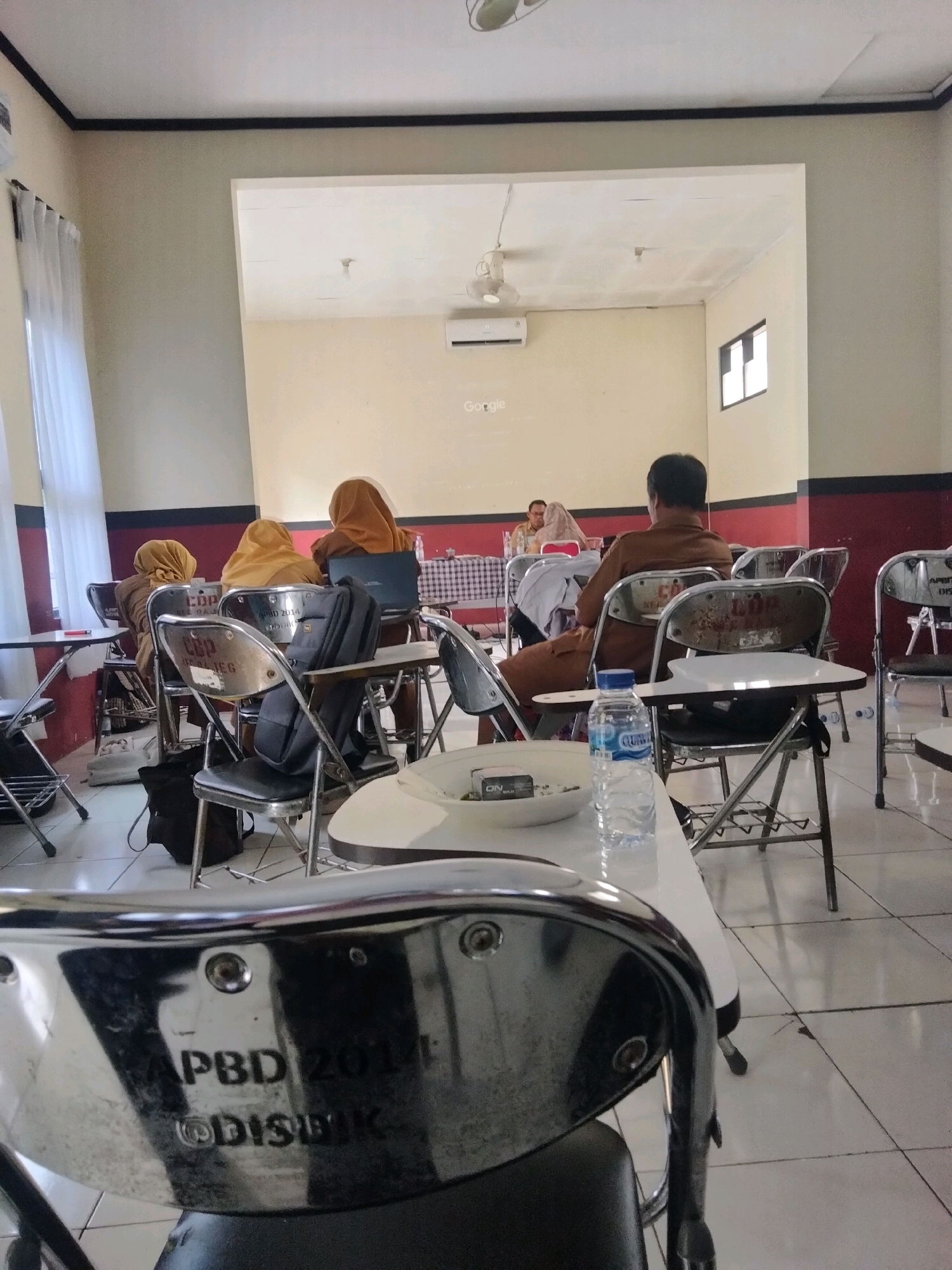 The image depicts an indoor scene, possibly a classroom or meeting room, with several people seated at desks. The environment appears to be sparsely decorated, with a projector screen visible on one wall. The people are primarily facing forward, suggesting they are engaged in a presentation or discussion. Some individuals are wearing headscarves, indicating a cultural context, and one person is dressed in what appears to be a uniform. The room is well-lit, with natural light coming from a window and artificial lighting from the ceiling. There are several chairs and tables, some of which have writing surfaces attached. A bottle of water and a plate with some items are visible on a table in the foreground. The overall impression is one of an educational or professional gathering. Without further information or context, it is not possible to definitively place the location within the city of Suka Tani, Indonesia.
