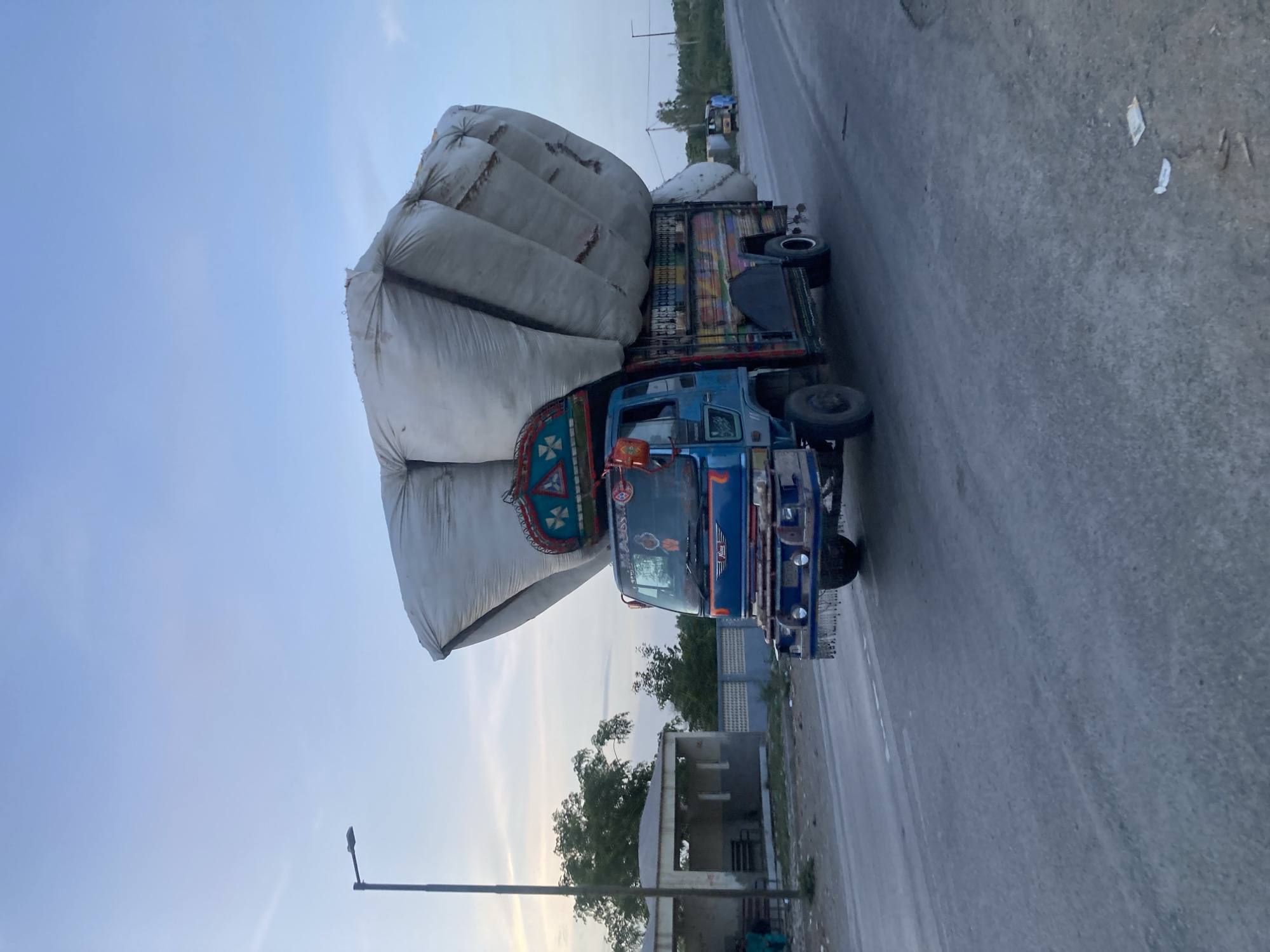 A truck heavily laden with large white bags is traveling on a paved road in Uthal, Pakistan. The truck is blue with colorful decorative elements. To the side of the road is a small building and some vegetation. A street light is also visible.