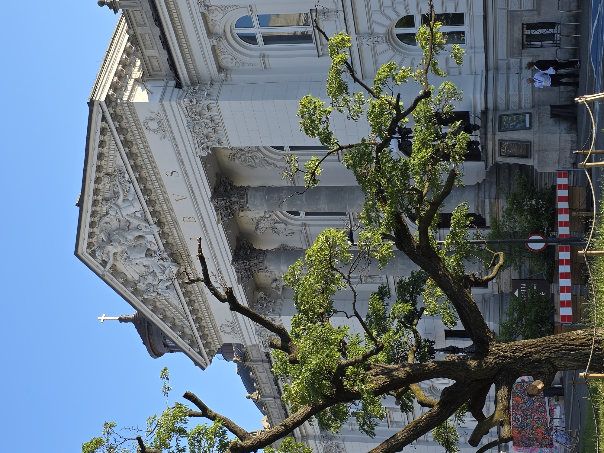 The image shows the facade of the Polish Academy of Sciences building, located at 72 Krakowskie Przedmieście in Warsaw, Poland. The building is made of white stone and features classical architectural elements such as columns, pediments, and ornate sculptures. A large, mature tree with green leaves partially obscures the left side of the building. The sky is clear and blue. In the foreground, there is a sidewalk with a few people and some temporary barriers. One person, a man in a white shirt and dark pants, is standing and looking to the right. Another person, a woman in a dark jacket and pants, is standing next to him, also looking to the right. They appear to be engaged in a conversation or waiting. The overall scene suggests a daytime setting with good weather.
